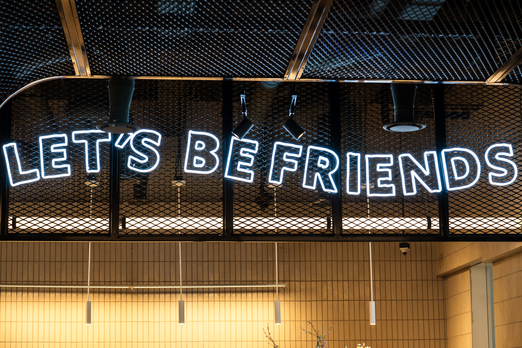 A neon sign reading "LET'S BE FRIENDS" is mounted on a mesh panel above a tiled wall in an indoor setting.