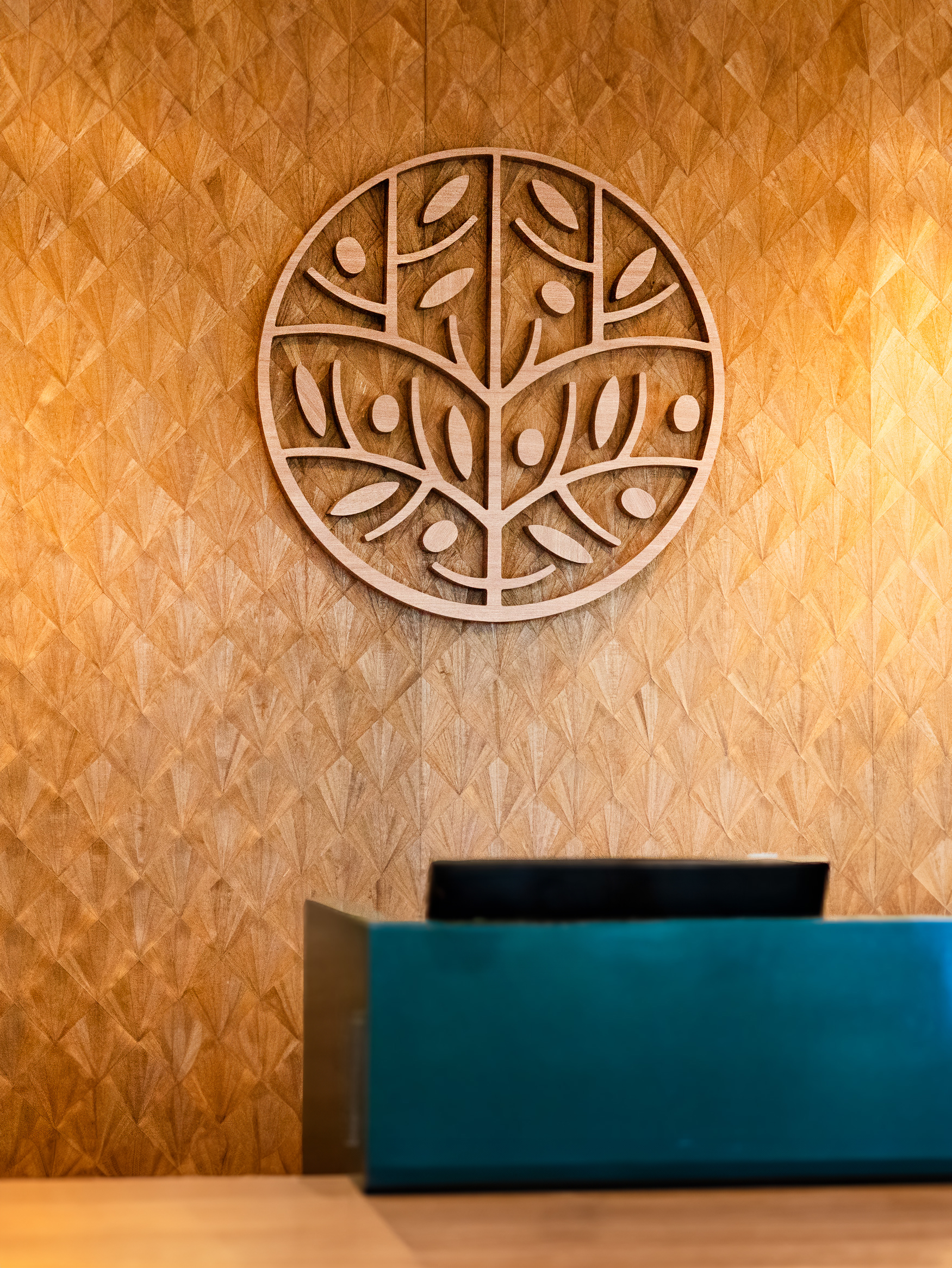 Reception desk with a black monitor in front of a wooden wall featuring a large circular tree and leaf design.