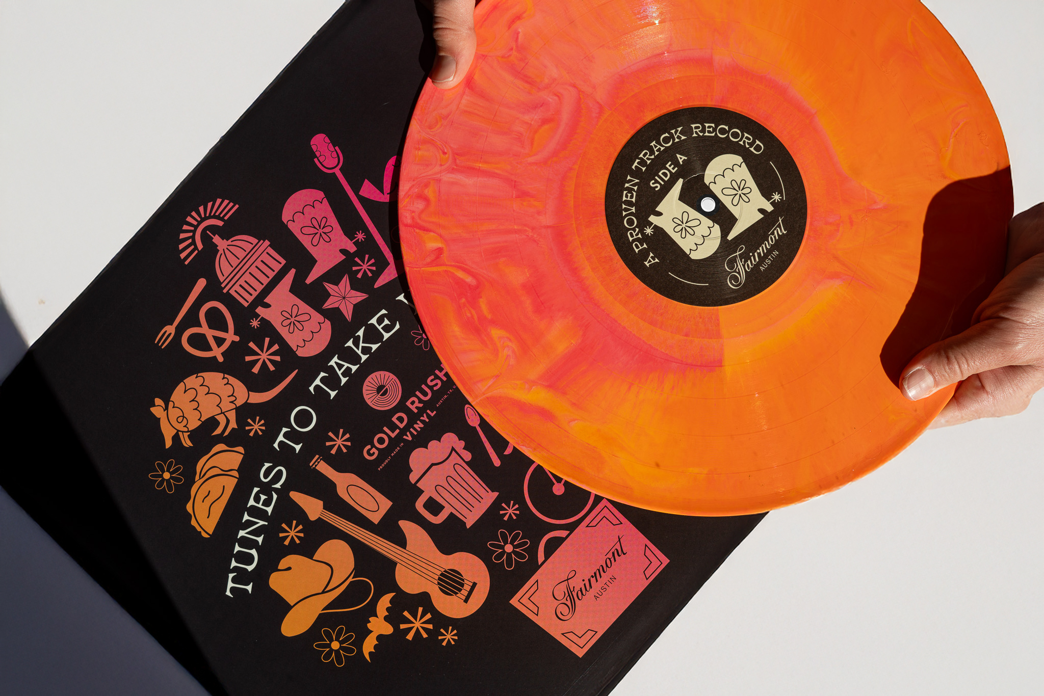 A person holds an orange marbled vinyl record above a black and pink "Tunes to Take" album cover featuring various illustrated icons and the Fairmont logo.