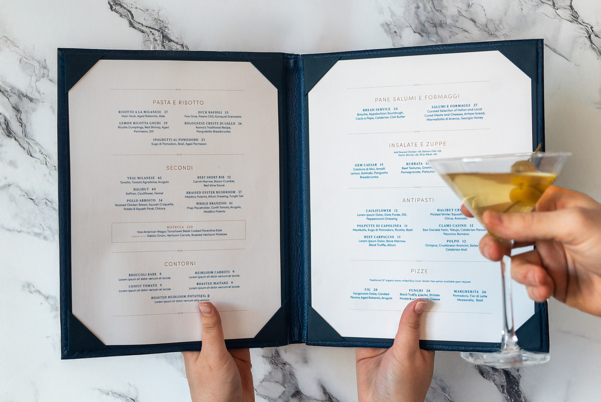 A person holds an open restaurant menu in one hand and a martini glass with an olive in the other; the menu has Italian food sections.