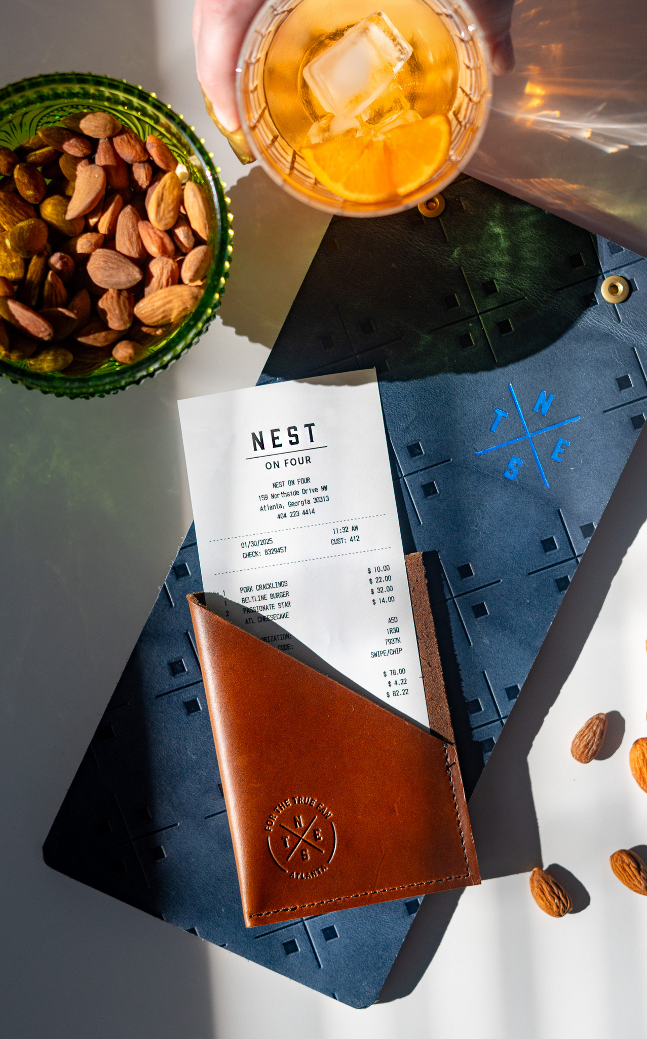 A restaurant receipt from Nest on Four sits in a brown leather holder on a blue placemat, next to a bowl of almonds and a hand holding an iced drink.