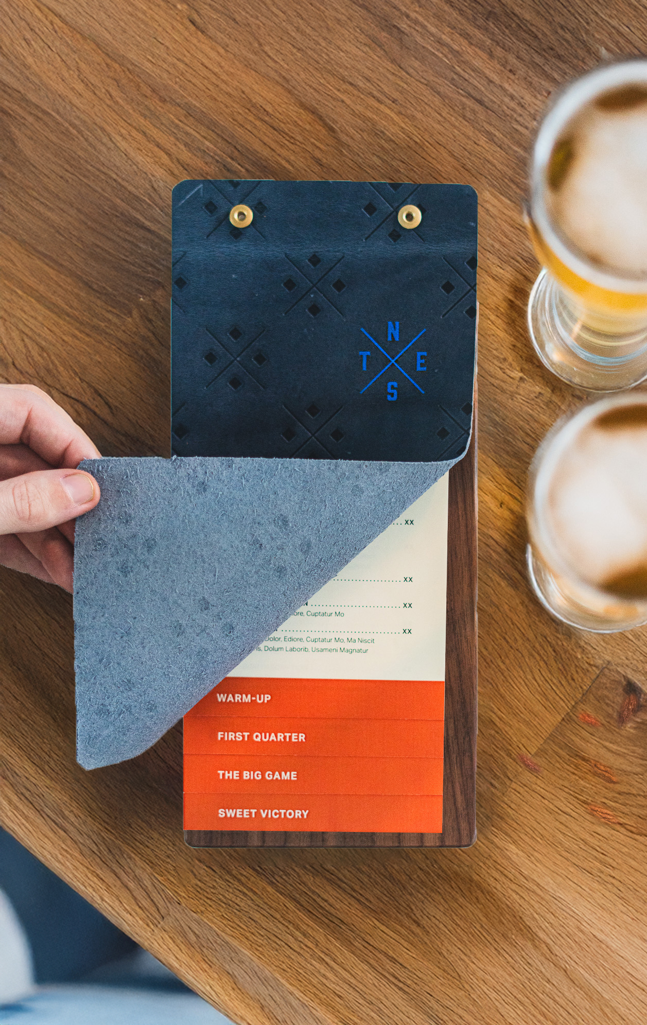 A person lifts a textured black cover from a menu with red and cream pages, next to two glasses of beer on a wooden table.
