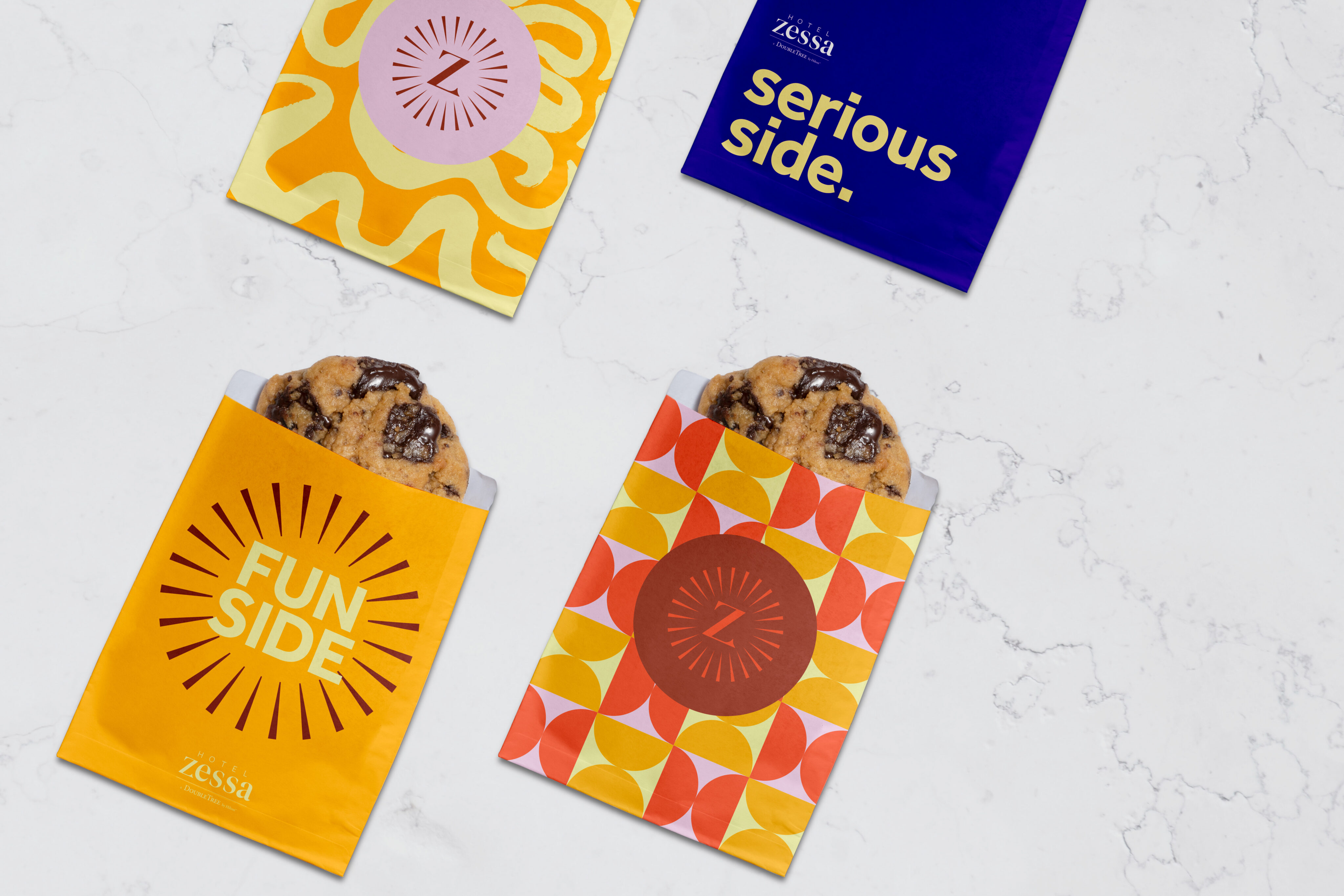 Four colorful cookie bags on a white surface; two bags are open showing chocolate chip cookies, and the others are sealed with graphic designs and text.