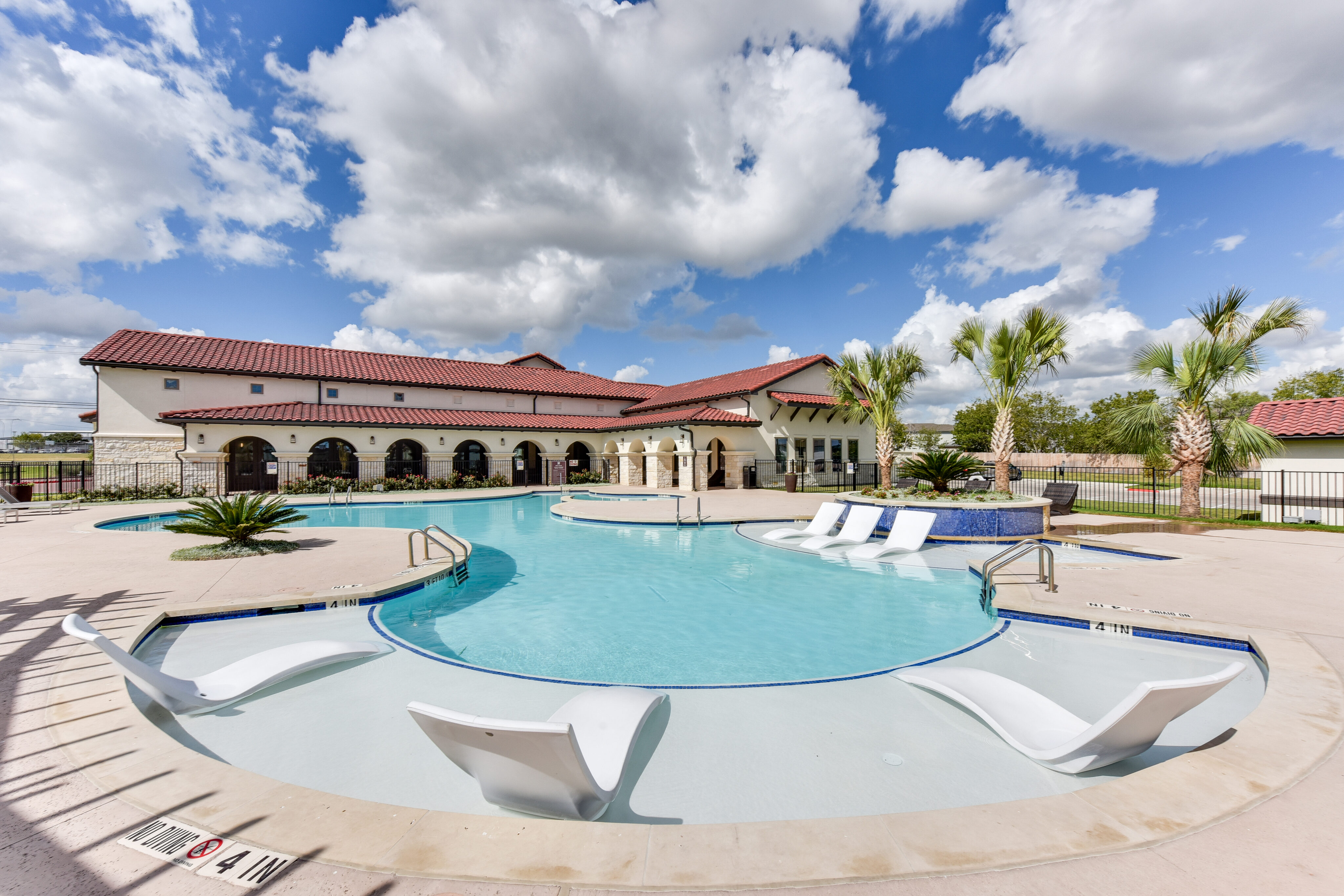 Outdoor swimming pool with built-in lounge chairs, a hot tub, and palm trees, in front of a large building with a red tile roof under a partly cloudy sky.
