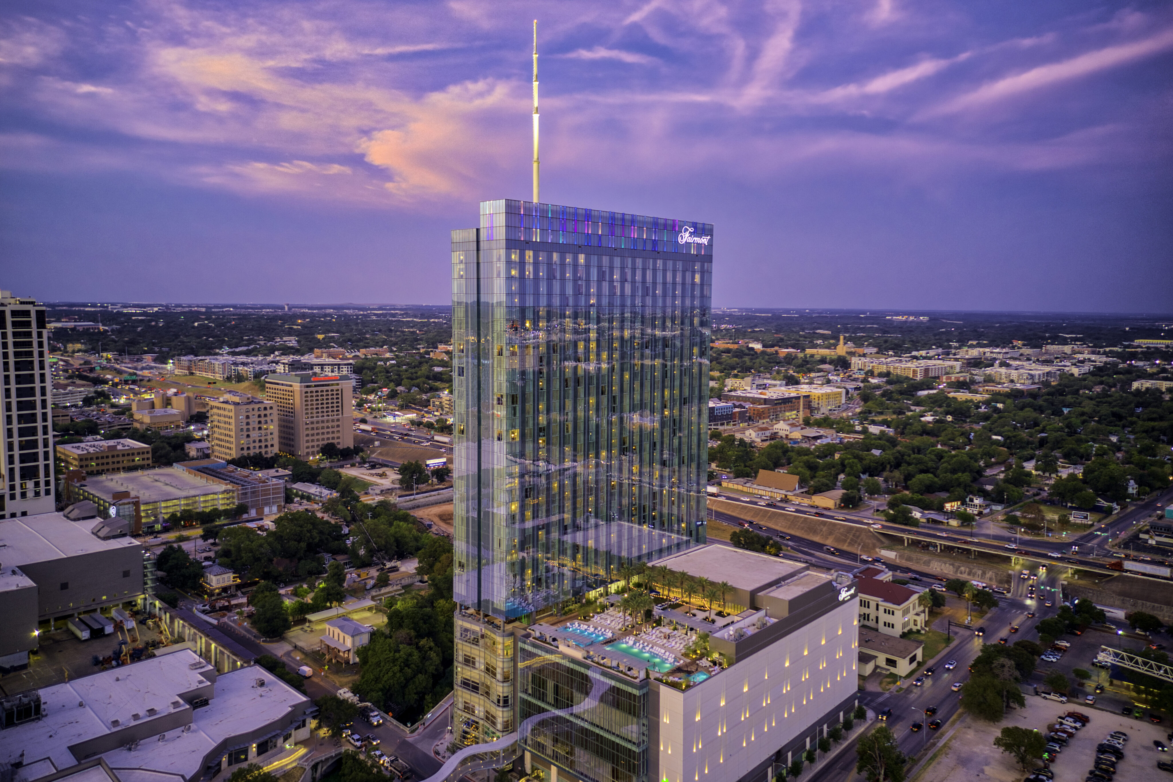 A tall, modern glass hotel building with a rooftop pool overlooks a cityscape under a colorful evening sky.