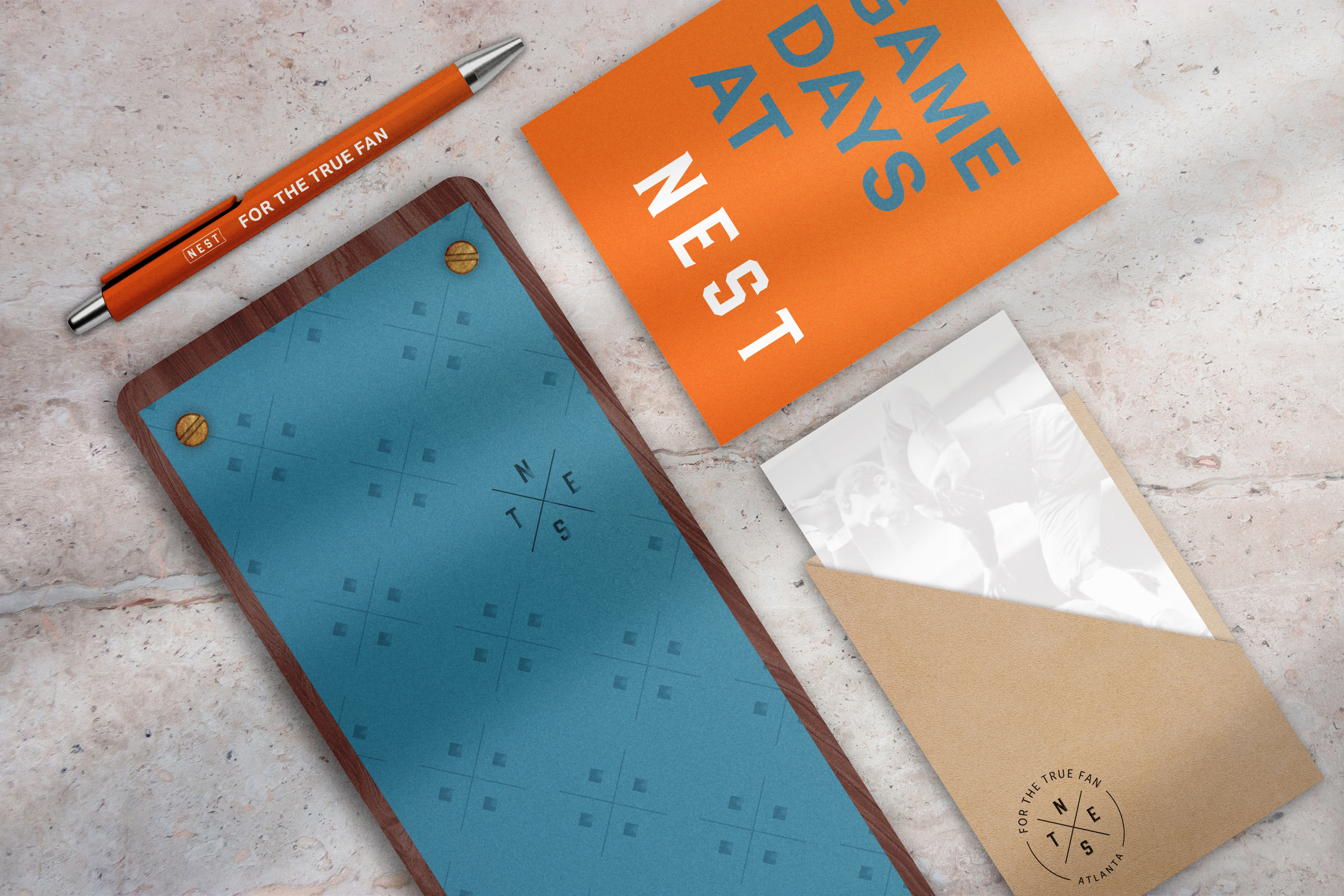 A pen, a blue notepad, an orange booklet labeled "GAME DAYS AT NEST," and a beige envelope with a photo and circular logo are arranged on a light stone surface.