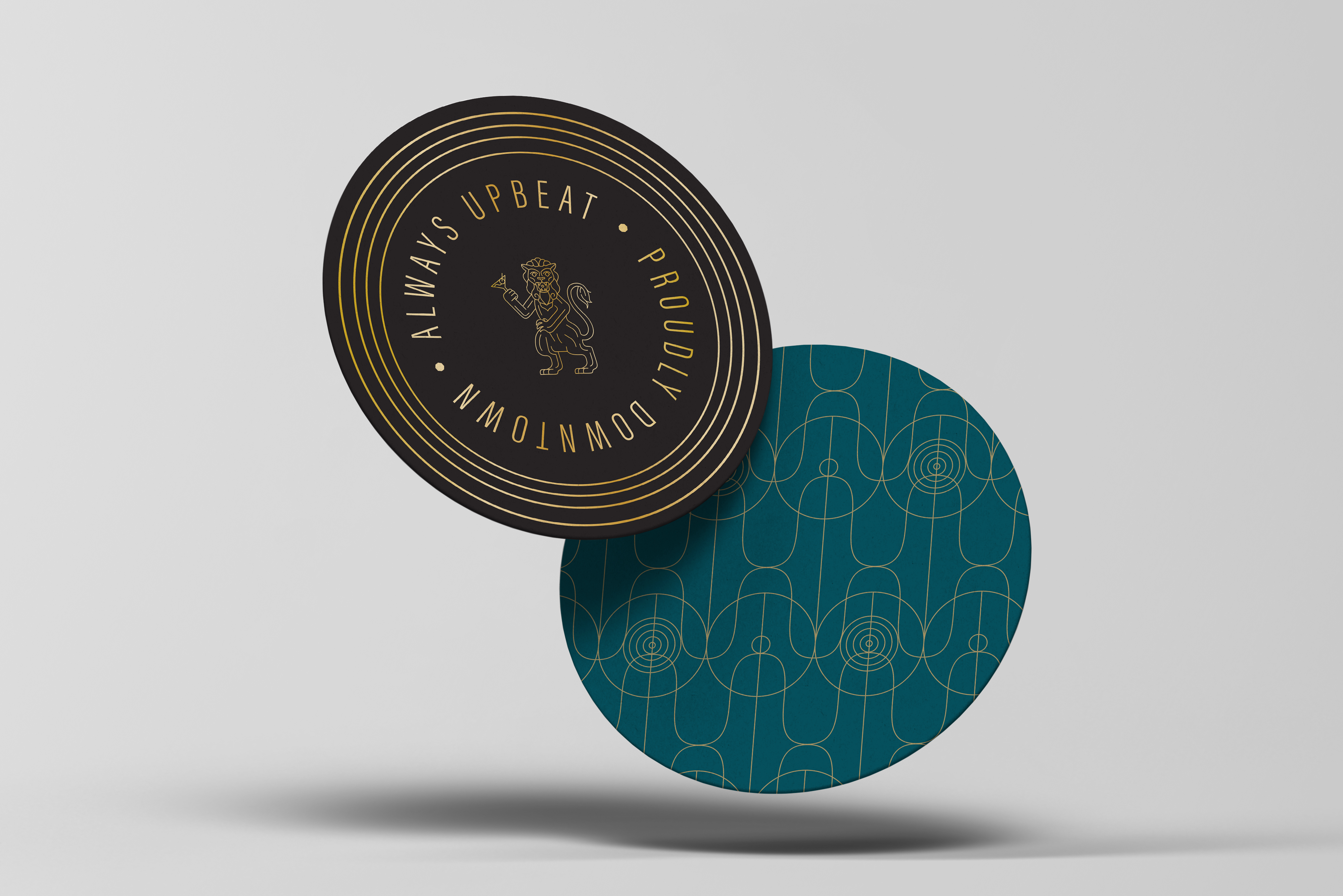 Two round coasters, one black with gold text and lion emblem reading "Always Upbeat • Proudly Downtown," and one teal with an abstract circular line pattern, are shown balancing together.