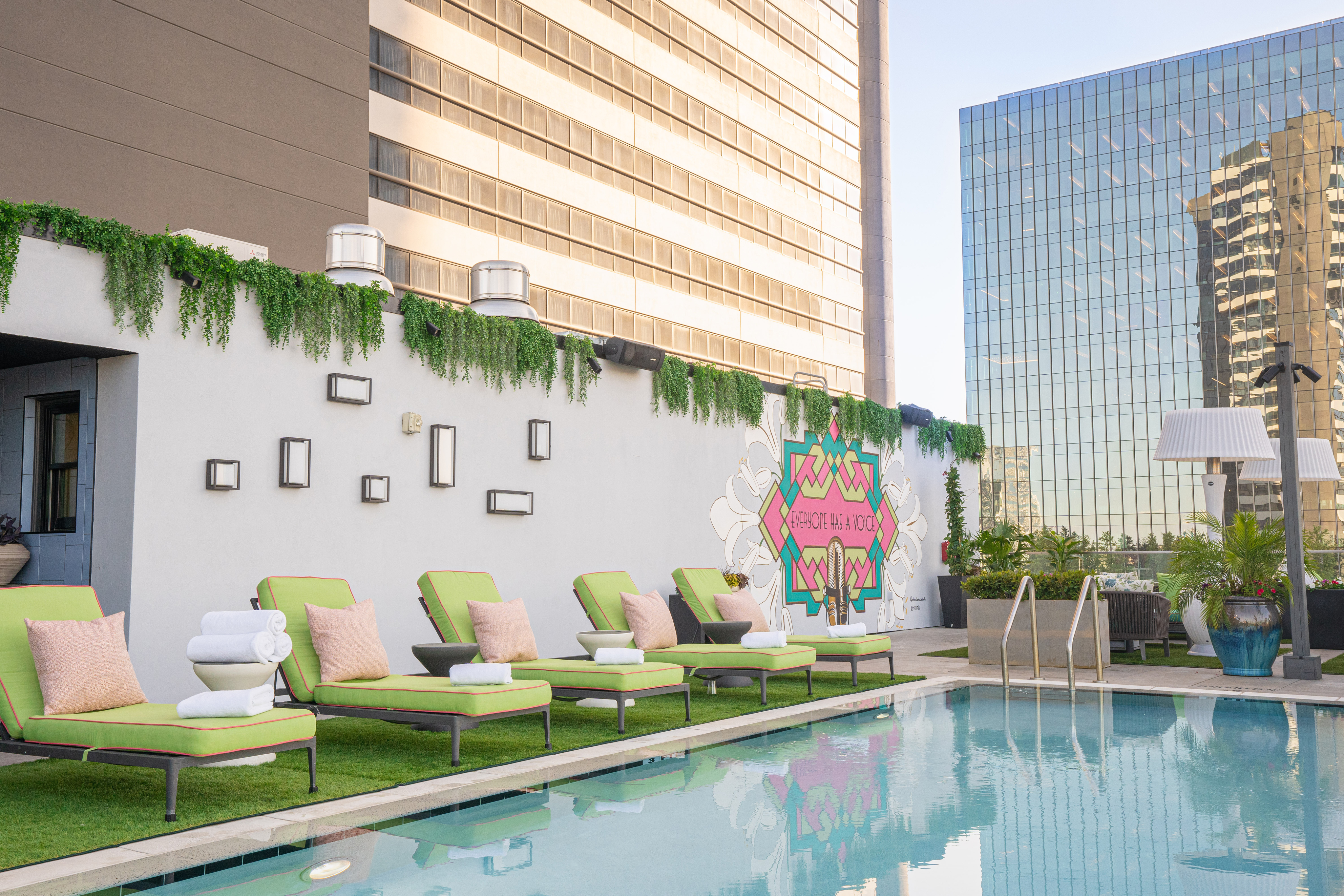 Rooftop pool with green lounge chairs, rolled towels, and a mural on a white wall, surrounded by tall modern buildings and greenery.