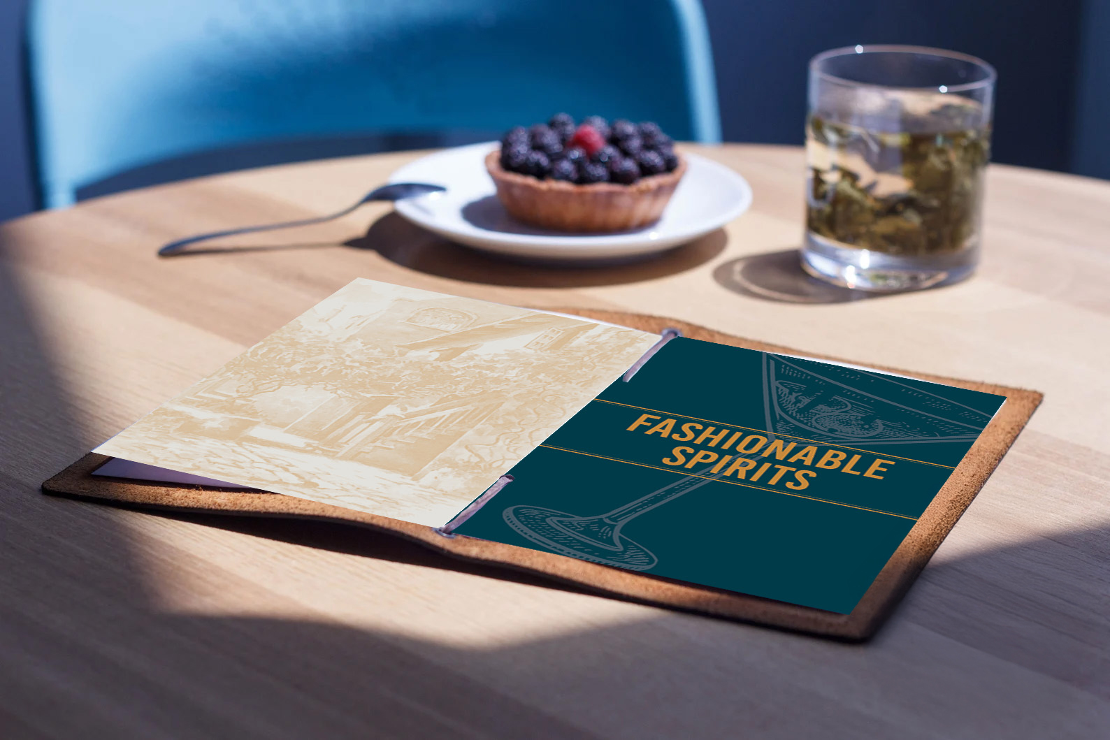 A leather menu labeled "Fashionable Spirits" on a wooden table, next to a berry tart and a glass of beverage, with a blue chair in the background.