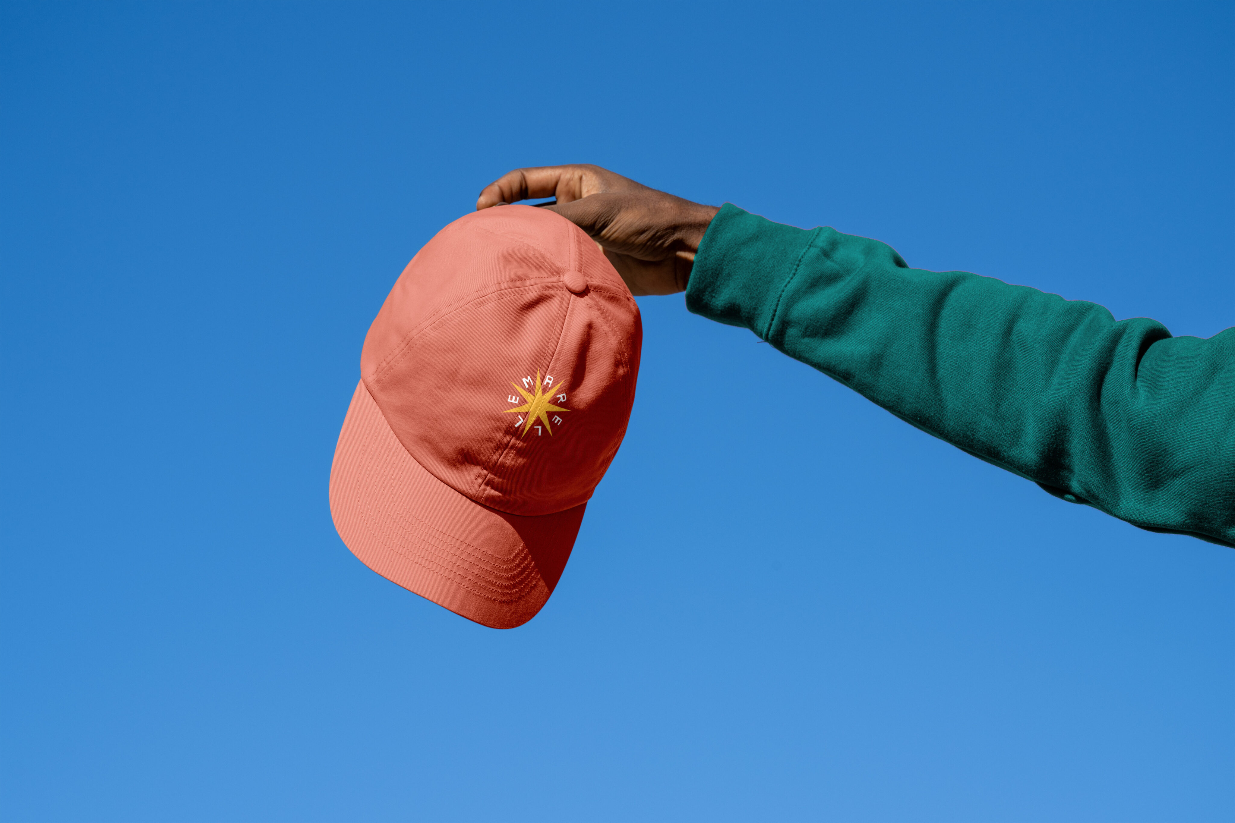 A person in a green sleeve holds up a coral-colored baseball cap with a small yellow design against a clear blue sky.