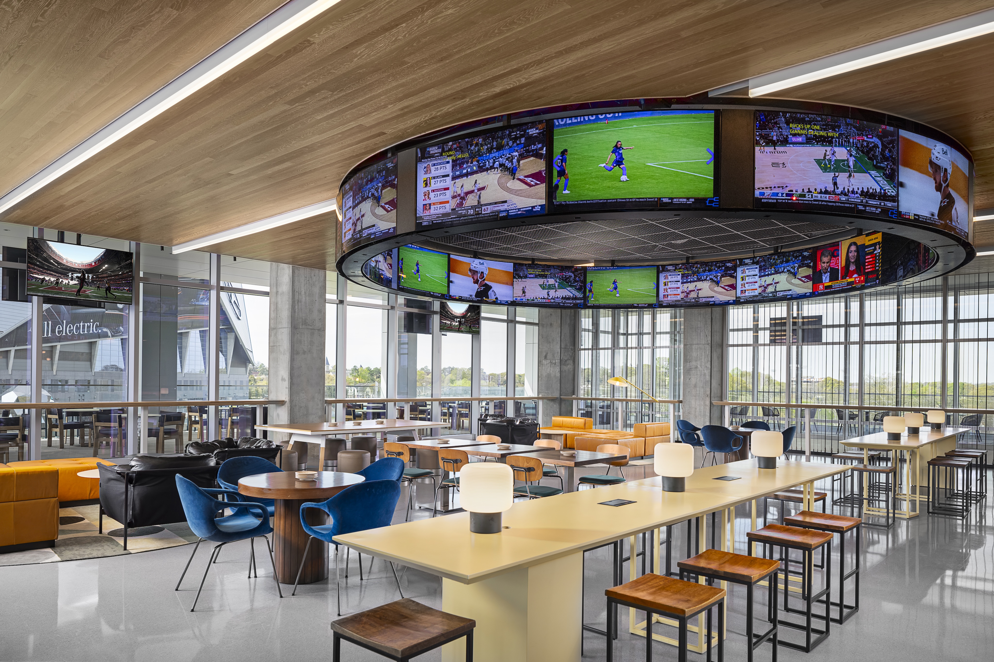 Modern lounge area with multiple tables, bar seating, and a circular overhead display showing various sports channels inside a building with large windows and natural light.