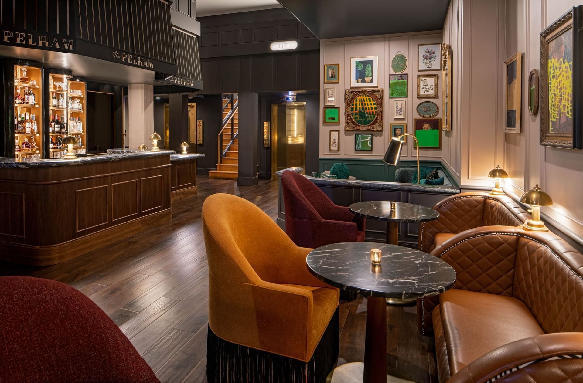 A stylish lounge with plush chairs, round black tables, a wooden bar, and eclectic framed art on the walls, featuring dark wood floors and soft lighting.