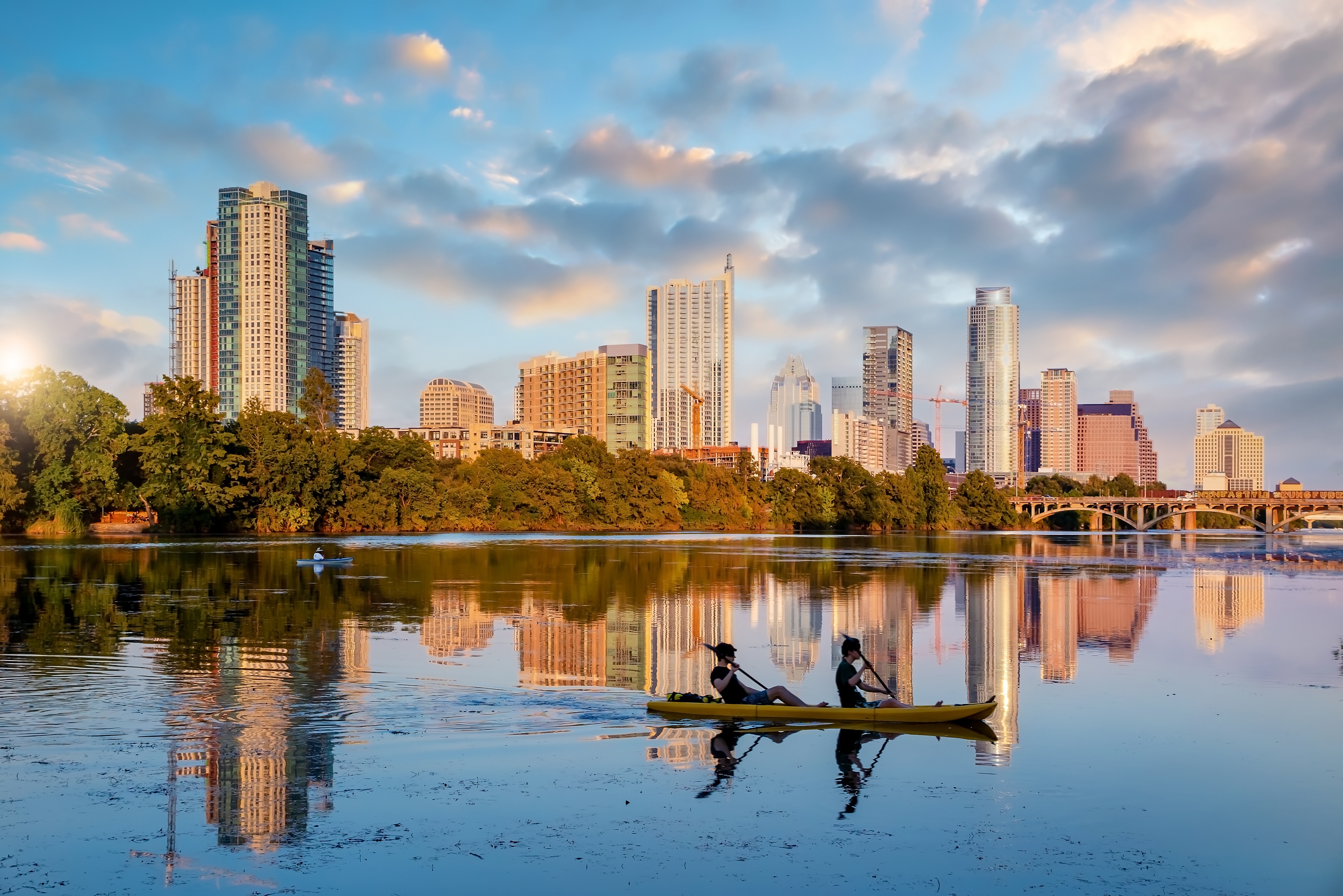 Two people paddle a yellow kayak on a calm river with a city skyline, trees, and a bridge in the background under a partly cloudy sky.