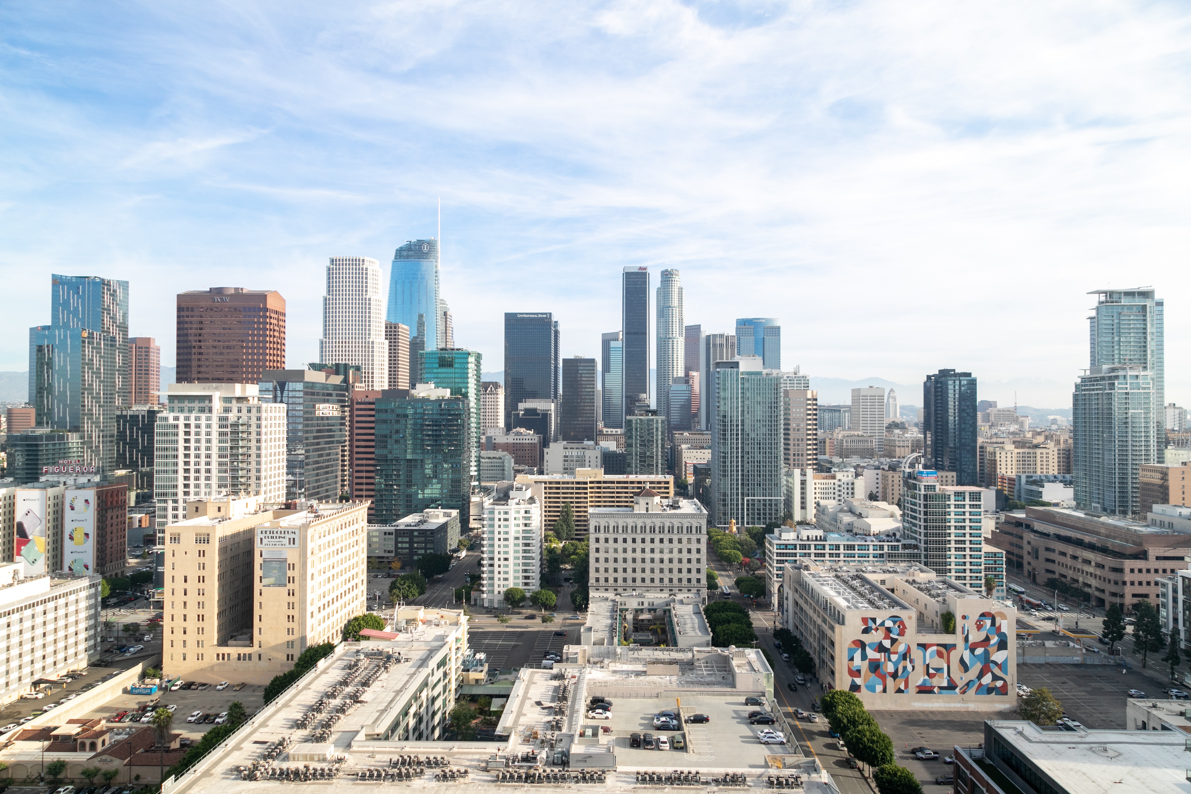 Aerial view of downtown Los Angeles featuring high-rise buildings under a partly cloudy sky, with various murals visible on lower buildings in the foreground.