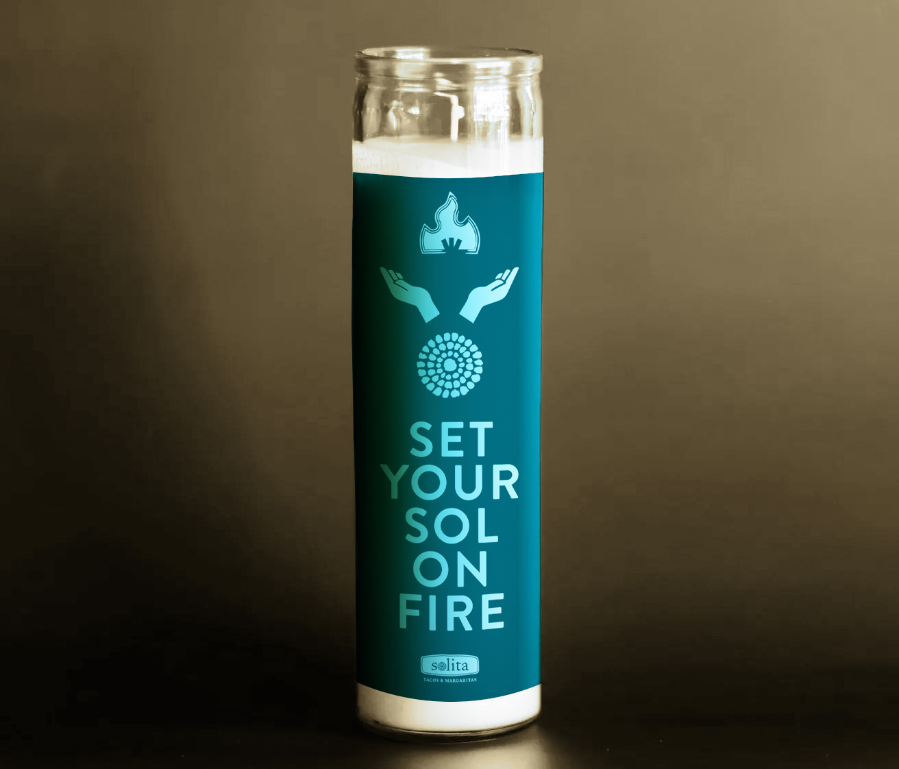 A tall glass candle with a teal label featuring hand and flame graphics and the text "SET YOUR SOL ON FIRE" in large letters.