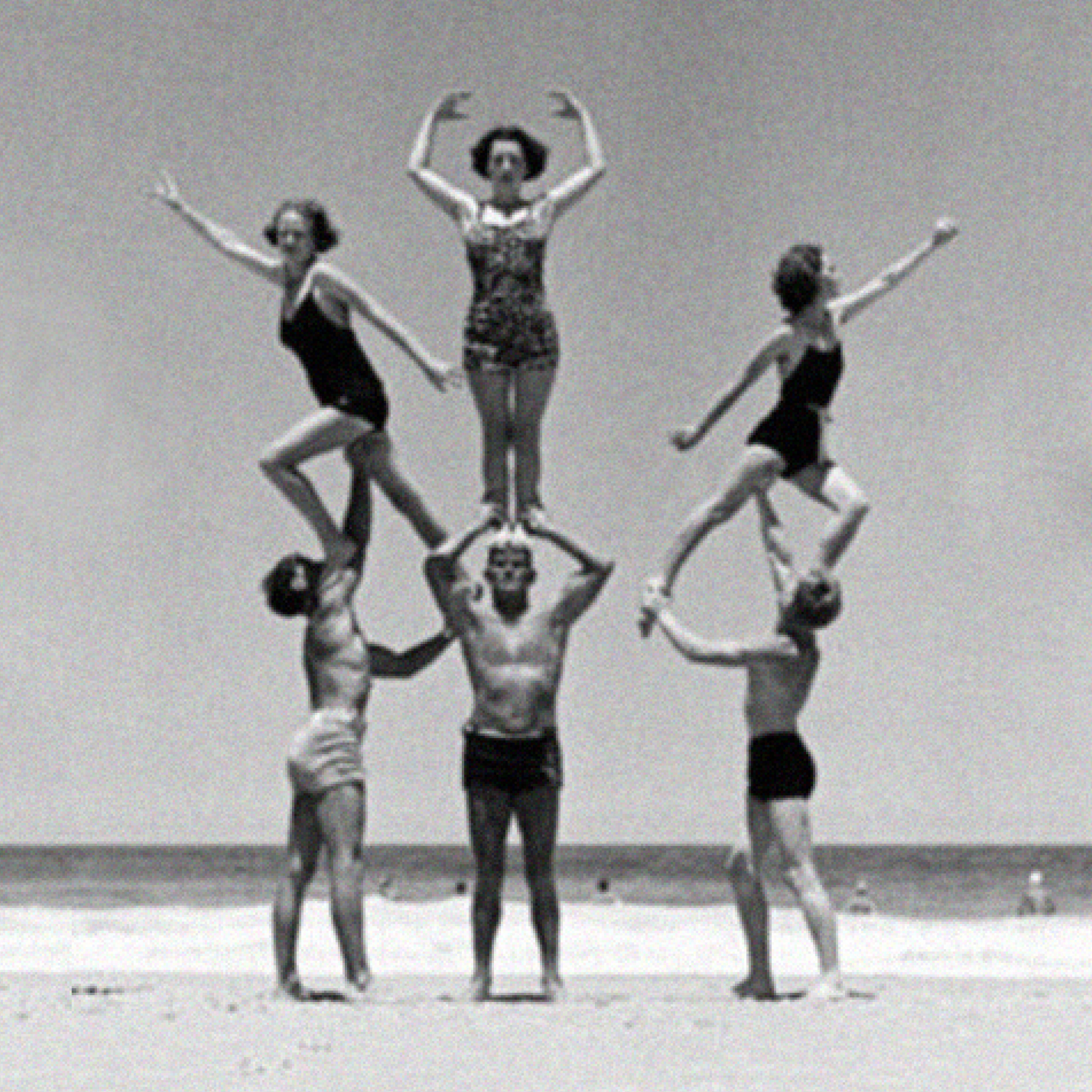 Three men in swimwear hold three women in a balanced human pyramid formation on a beach, with the ocean and sky in the background.
