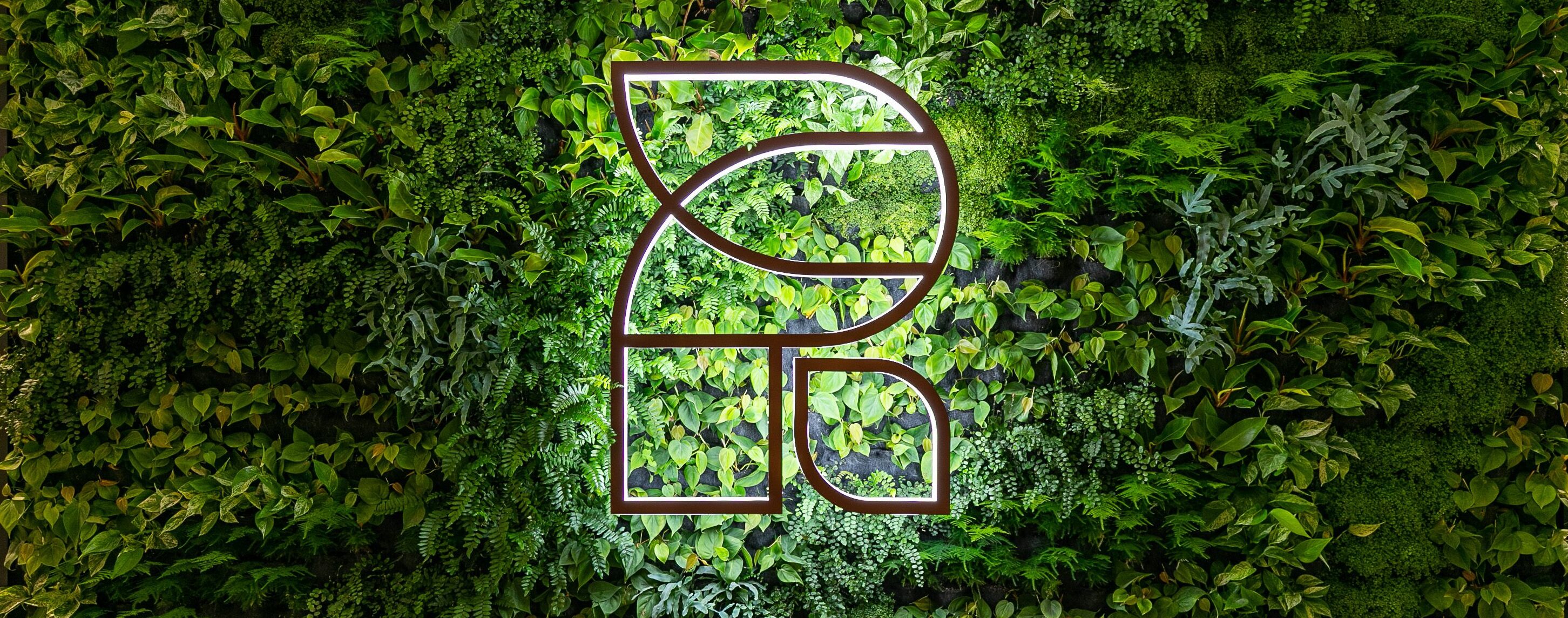 A green living wall with various plants and a large illuminated geometric logo in the center, set in a modern indoor space with wooden flooring.