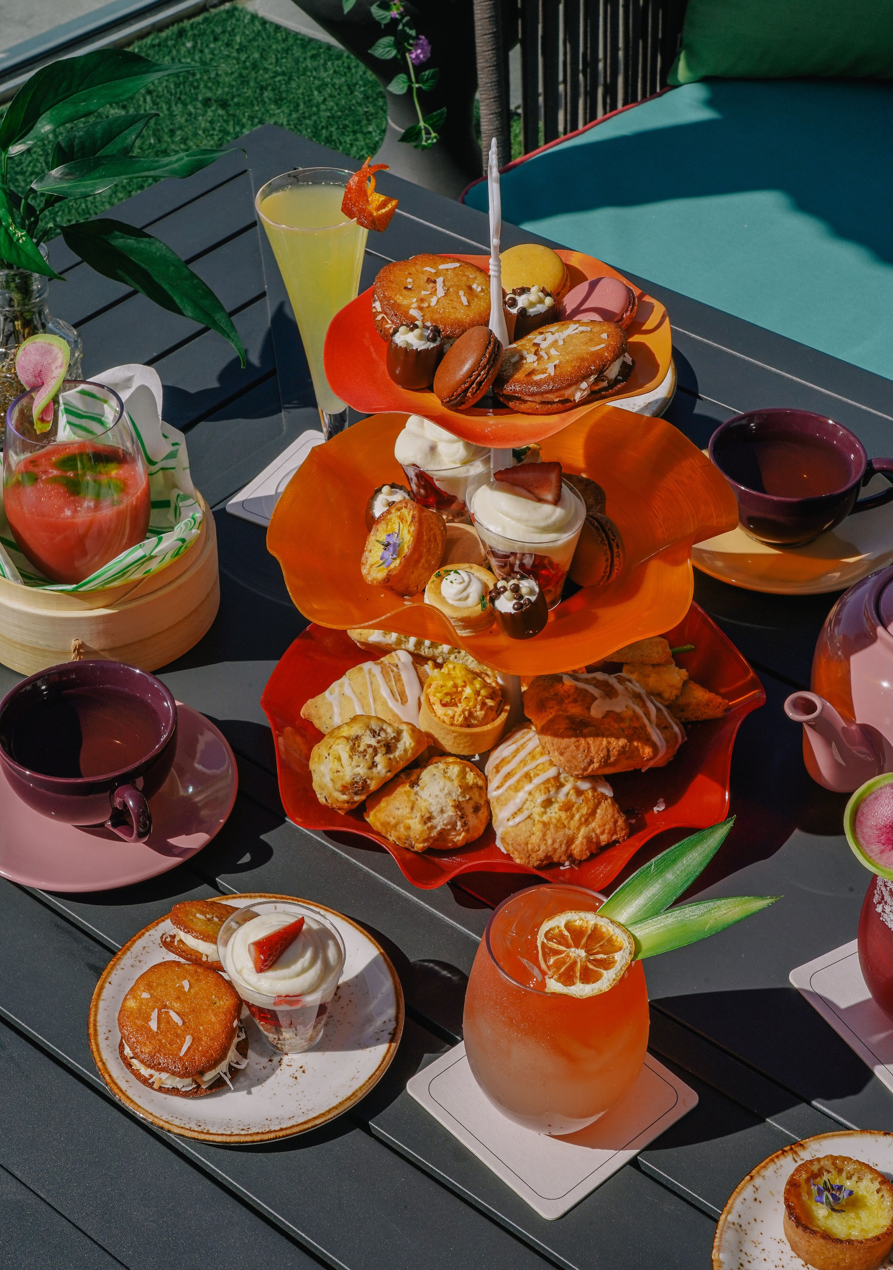 A three-tiered tray with assorted pastries and desserts is set on a table, surrounded by colorful drinks and teacups in an outdoor setting.