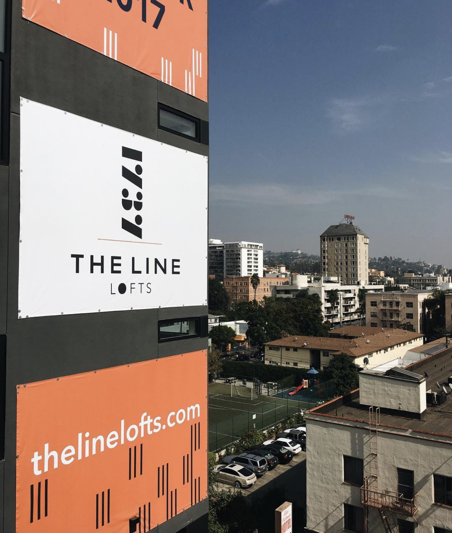 A building with "The Line Lofts" sign and website is shown in the foreground; city buildings and a soccer field are visible in the background under a clear sky.