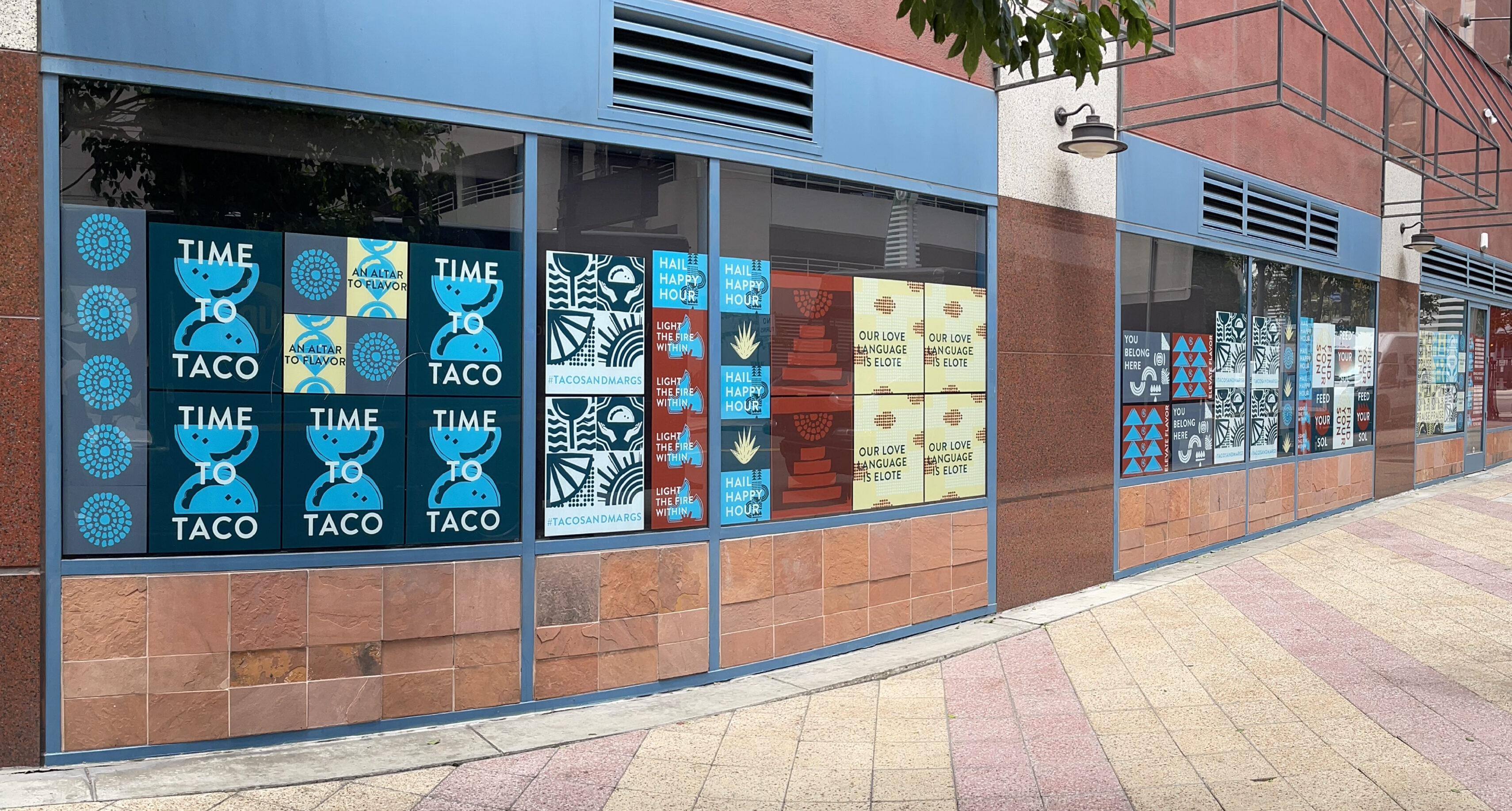 Storefront windows display multiple colorful posters, mainly blue and white, with taco-themed graphics and text such as "TIME TACO," along a paved city sidewalk.