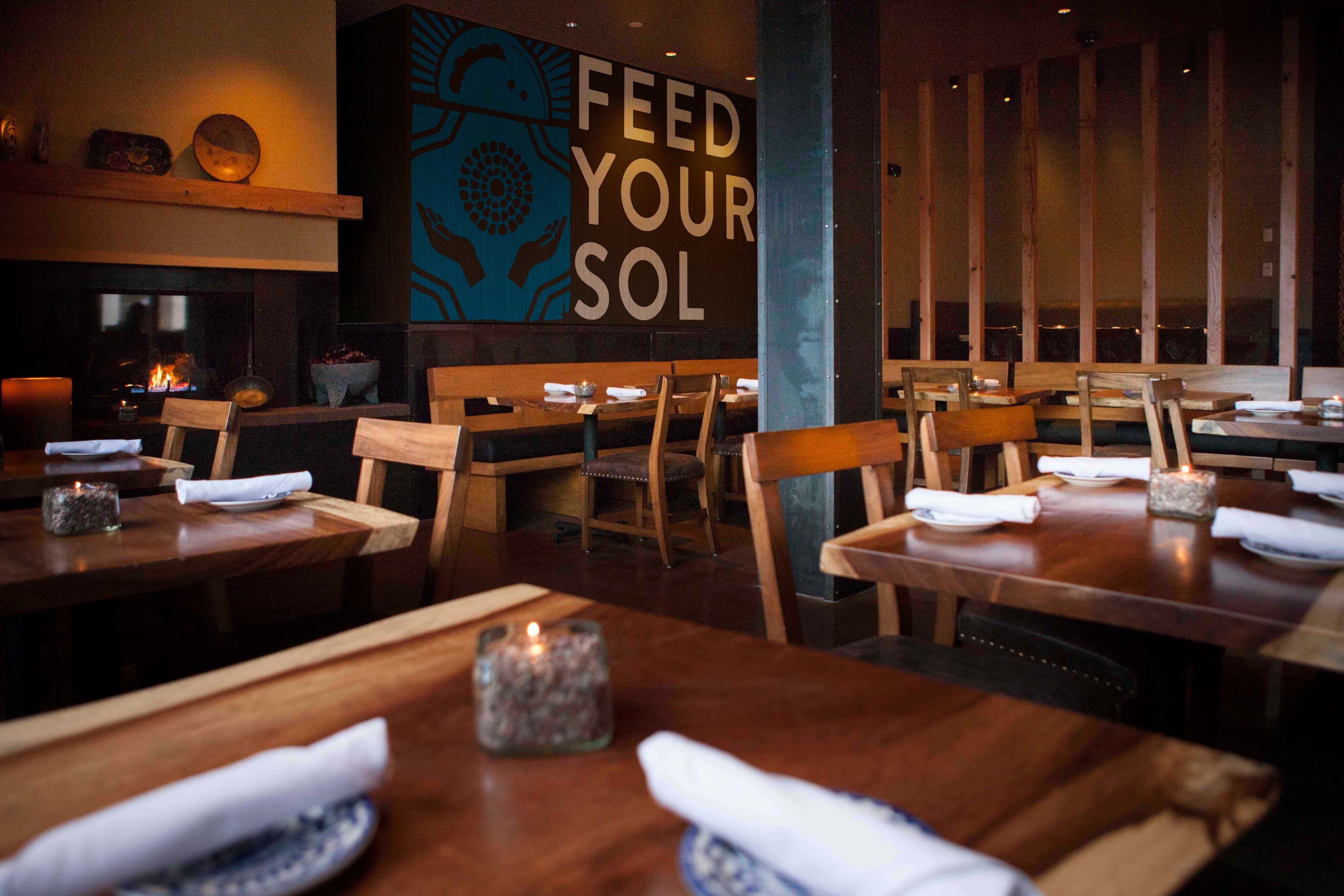 A modern restaurant dining area with wooden tables and chairs, set with plates and napkins; a wall mural reads "FEED YOUR SOL.