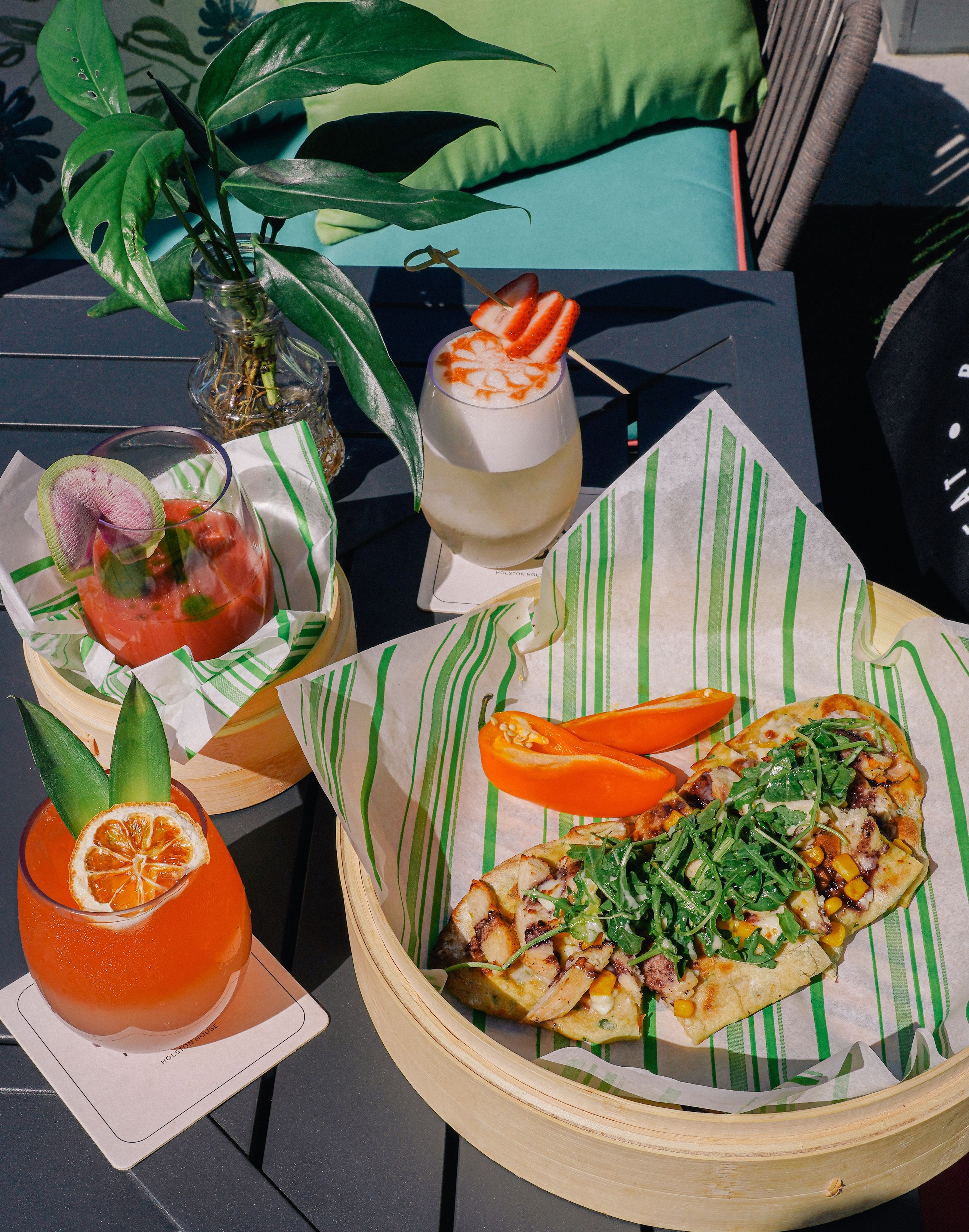 A table with three colorful cocktails and a flatbread topped with arugula, grilled chicken, corn, and two sliced orange peppers on striped paper. Decorative leaves in a glass vase.