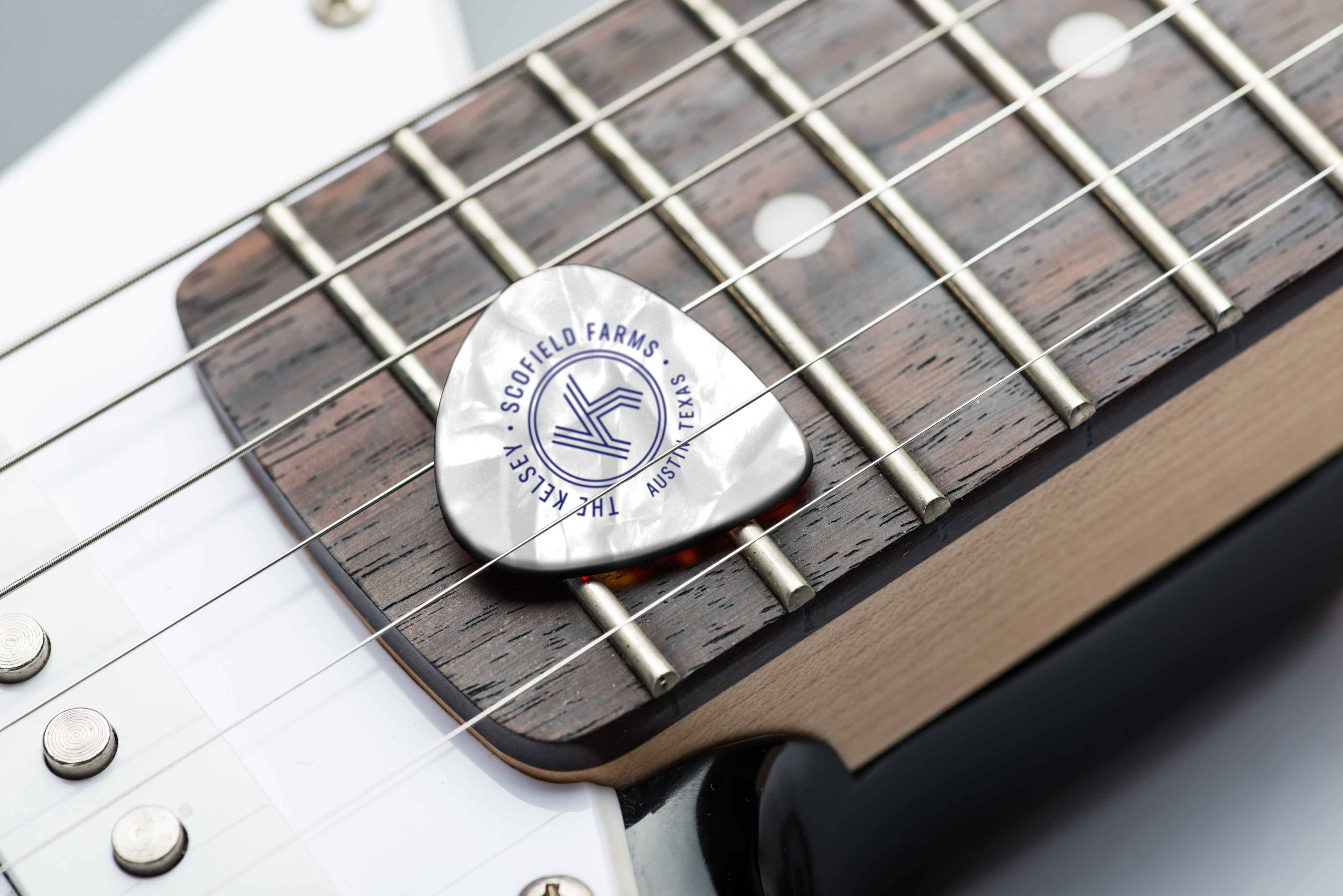 A metallic guitar pick with "Scofield Farms, Texas, The Keys" logo rests on the strings and fretboard of an electric guitar.