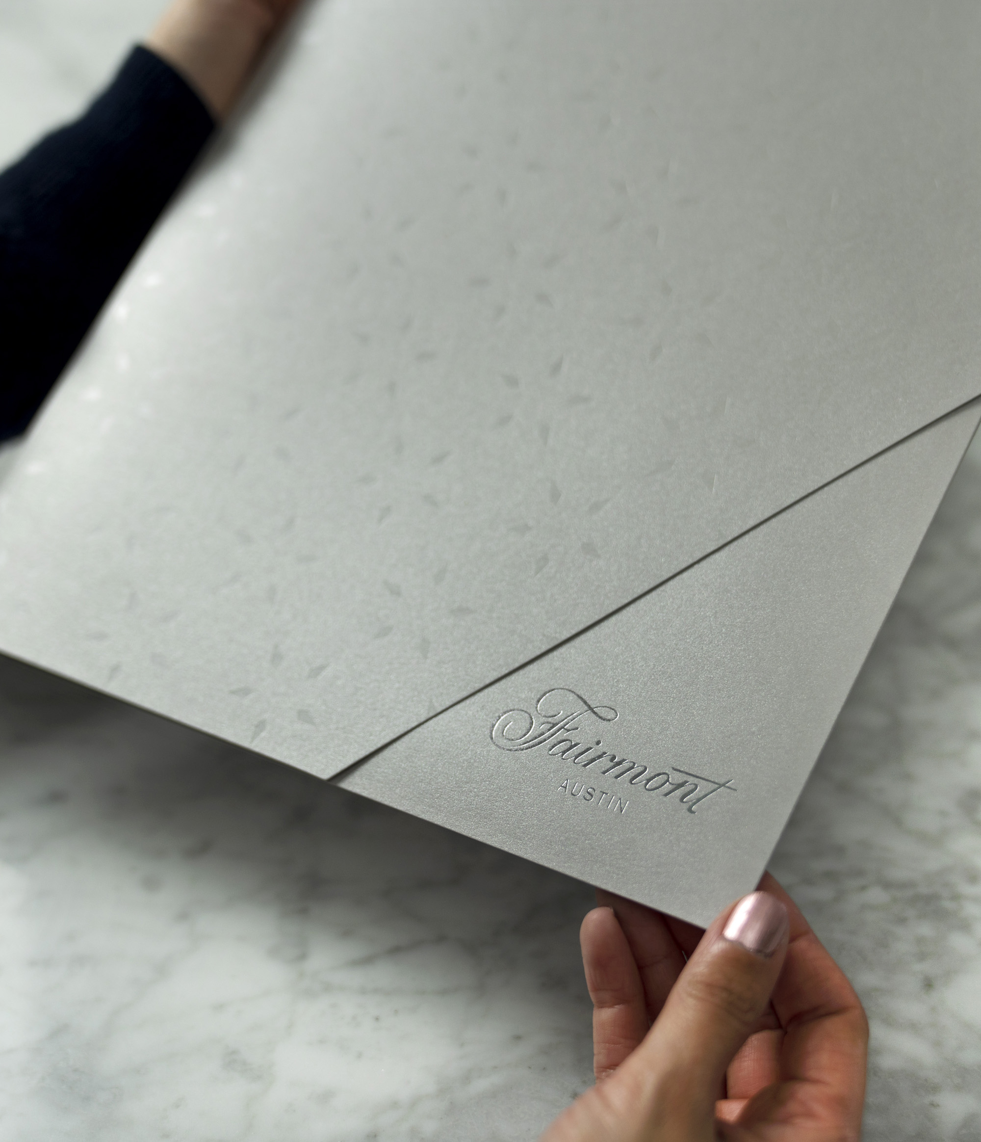 A person holds a large, silver folder embossed with “Fairmont Austin” on a marble surface.