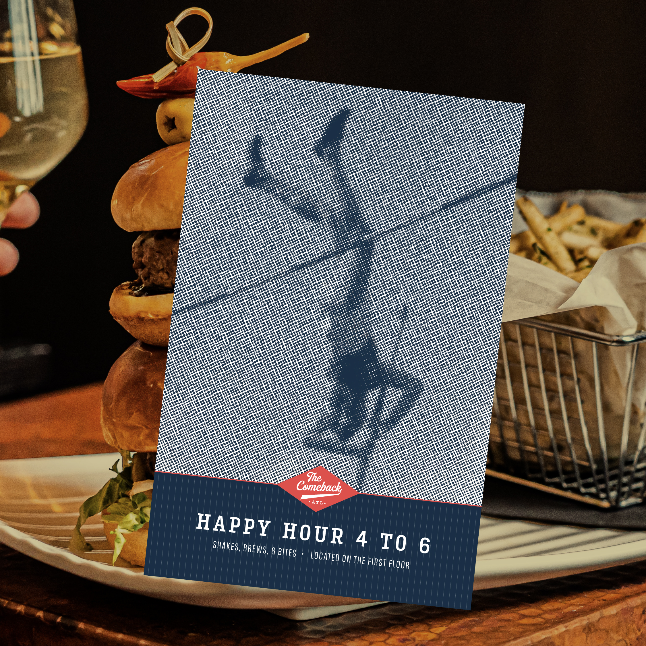 A happy hour promotional card stands in front of a plate of burgers and fries, with a person holding a wine glass in the background.