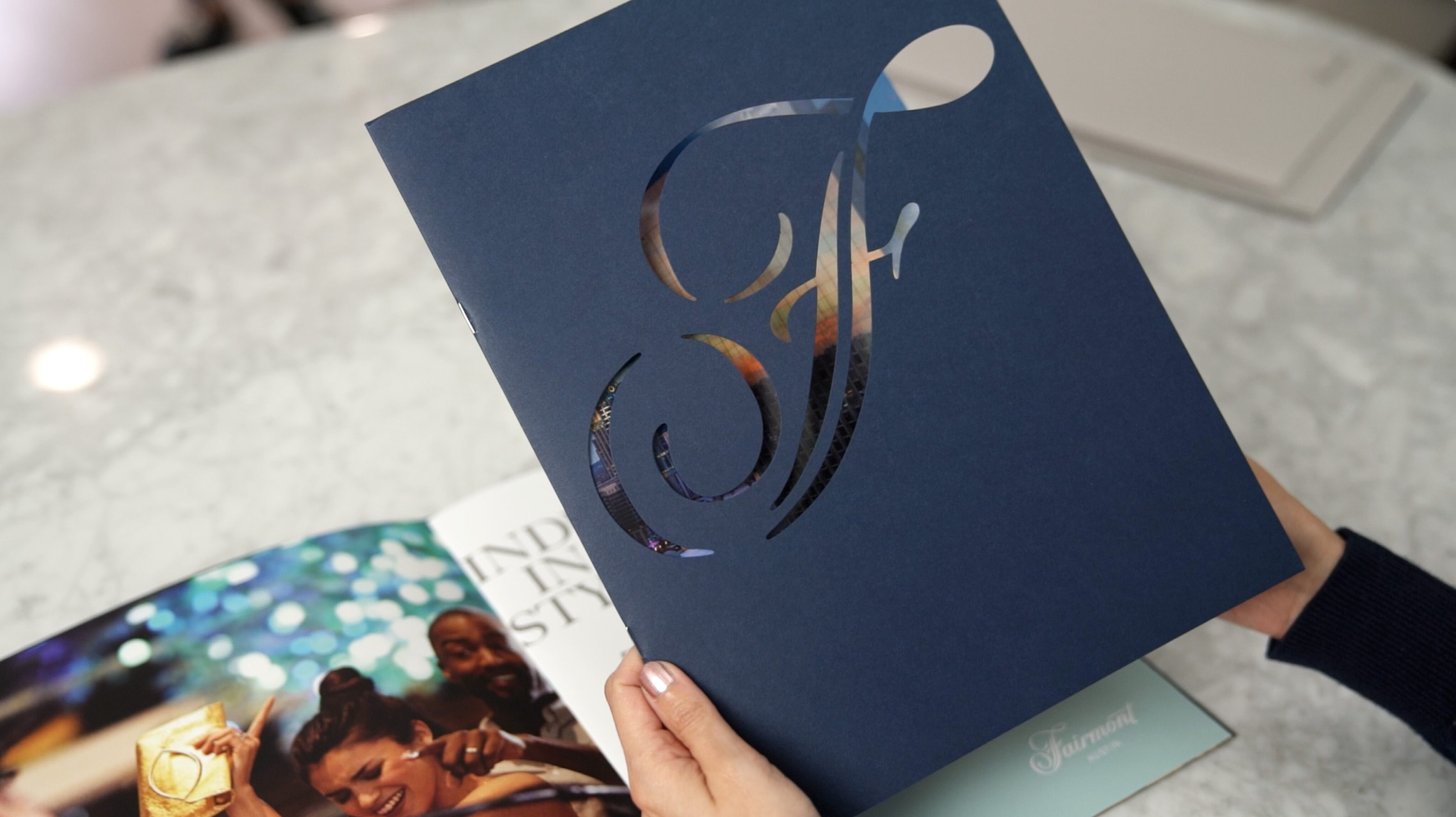A person holds a navy blue folder with a decorative cutout design, showing a colorful image beneath; an open brochure lies on the table nearby.