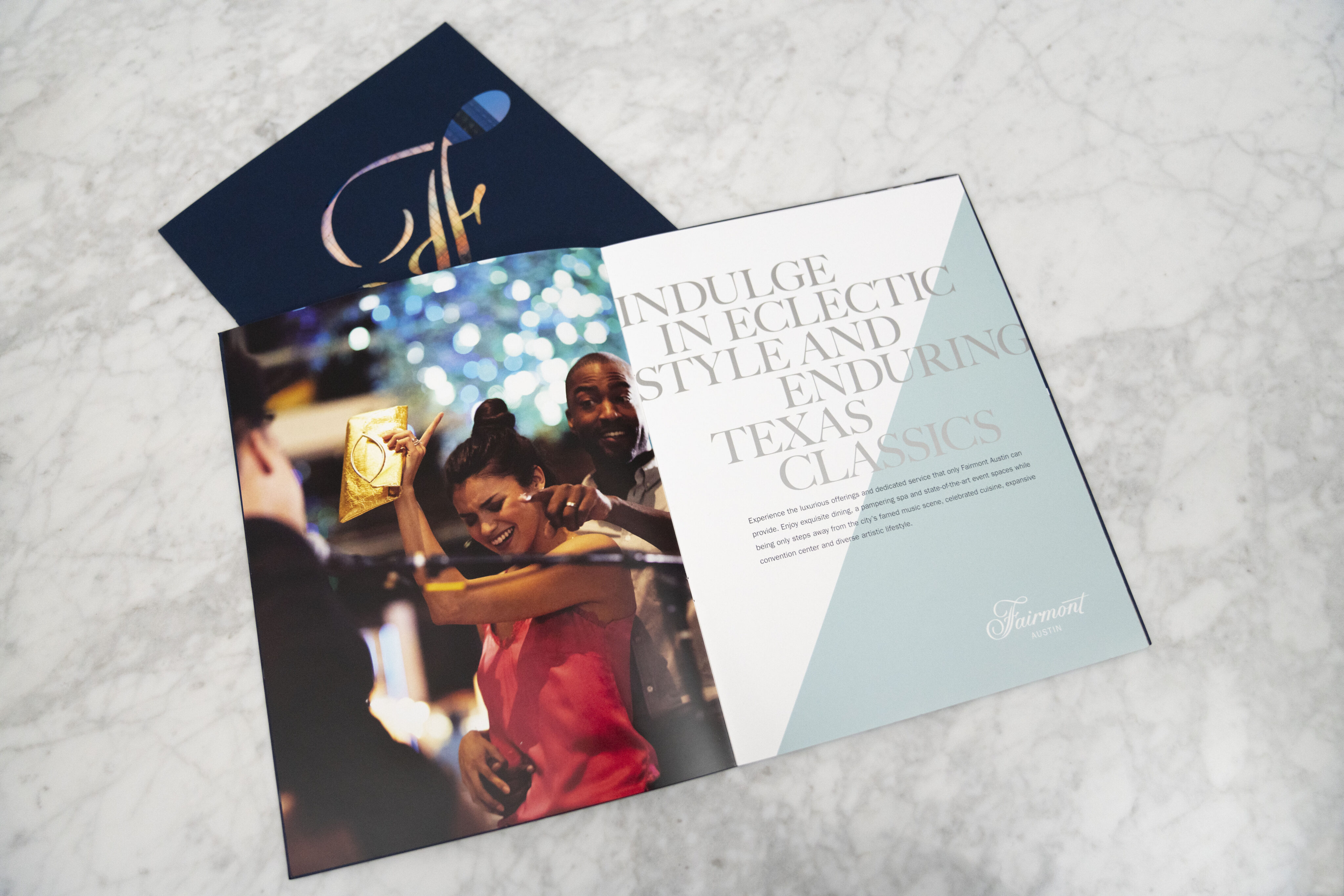 An open brochure on a marble surface displays a photo of people dancing and text reading, "Indulge in eclectic style and enduring Texas classics," with a Fairmont logo.