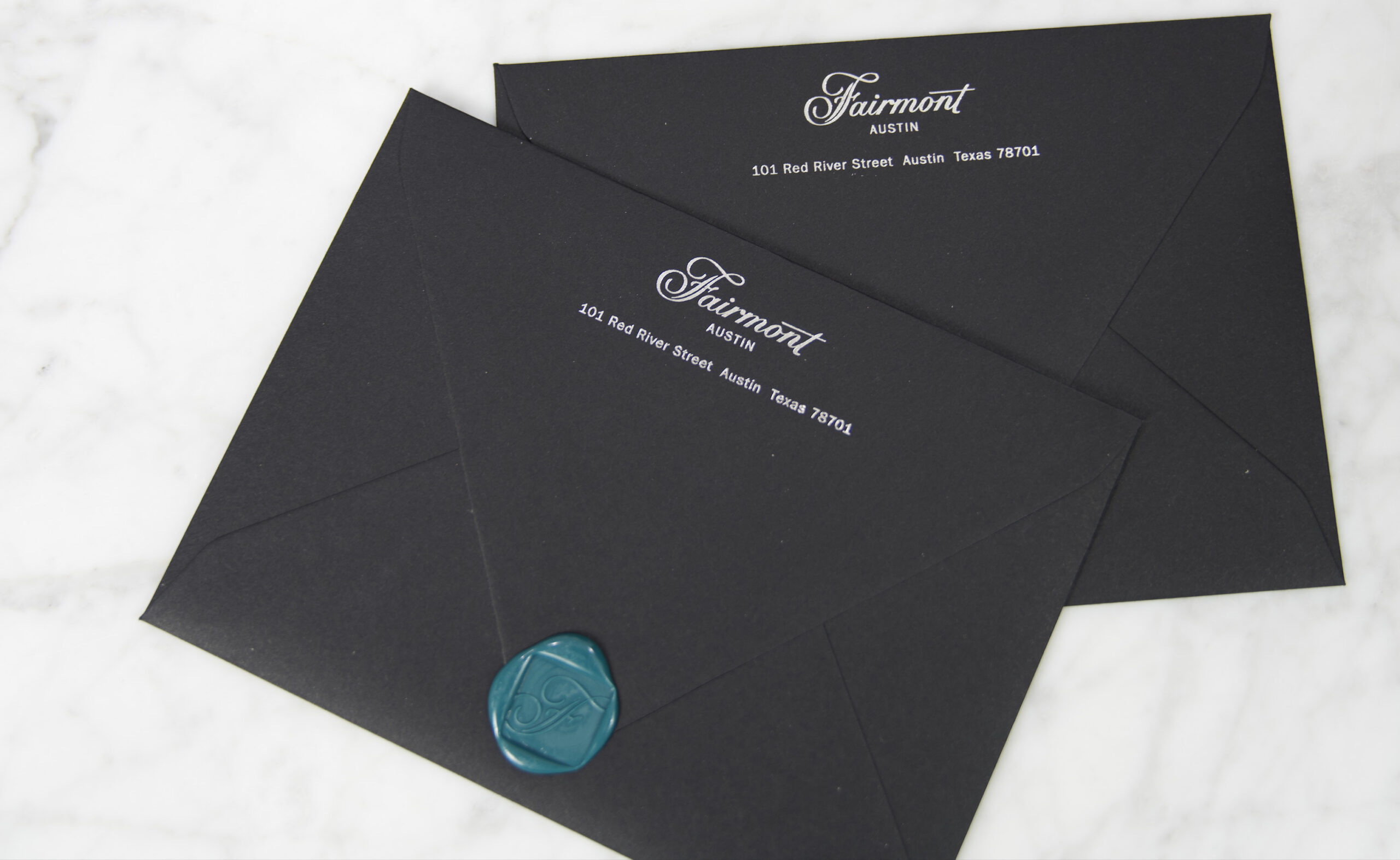 Two black envelopes with "Fairmont Austin" and an address printed in white; one envelope is sealed with a teal wax seal bearing an "F.