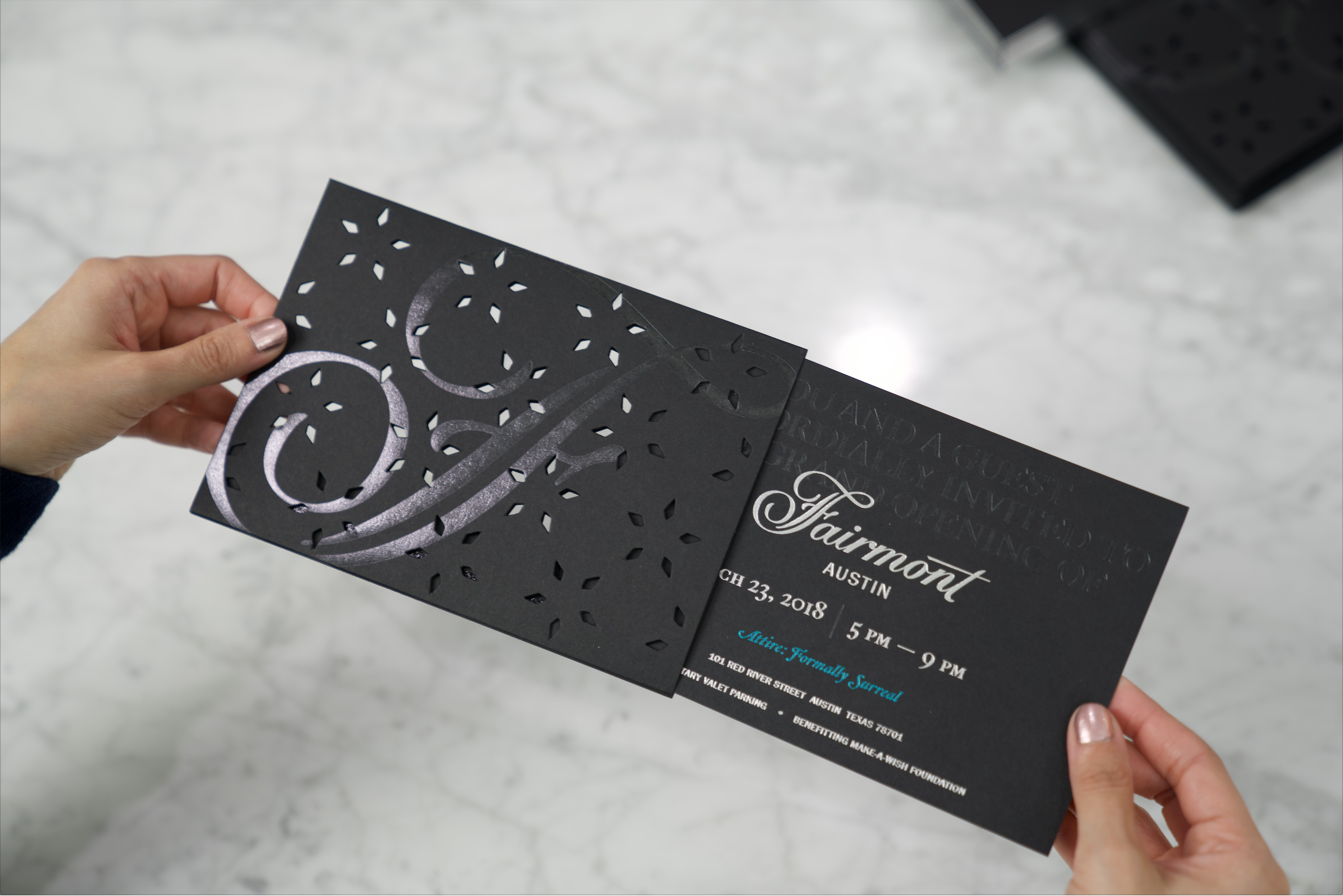 Person holding an elegant black event invitation card with decorative cut-out patterns and silver lettering, partially removed from its sleeve.