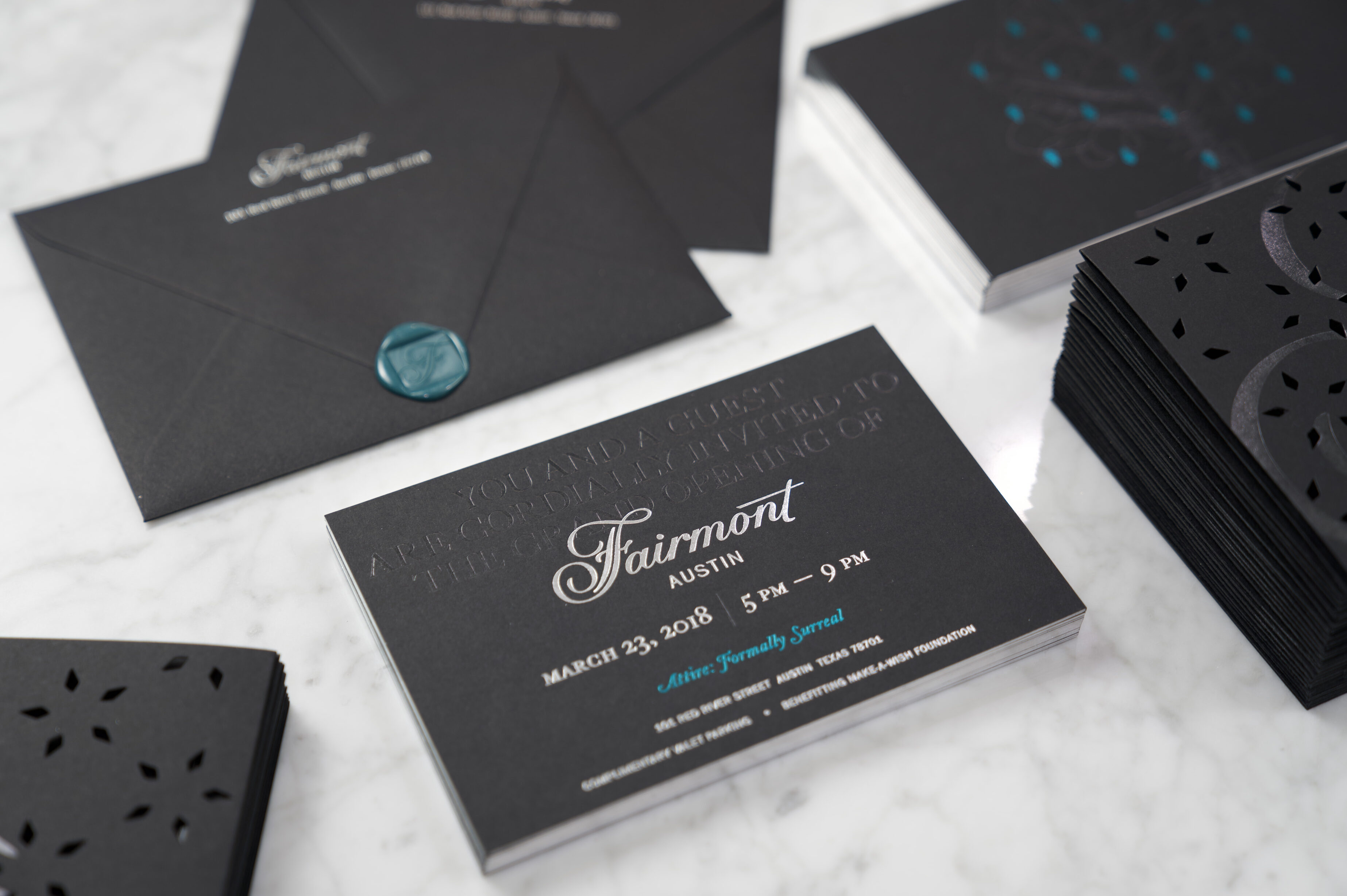 A black invitation card for Fairmont Austin's grand opening on March 23, 2018, is displayed on a marble surface with matching envelopes and decorative cutouts.