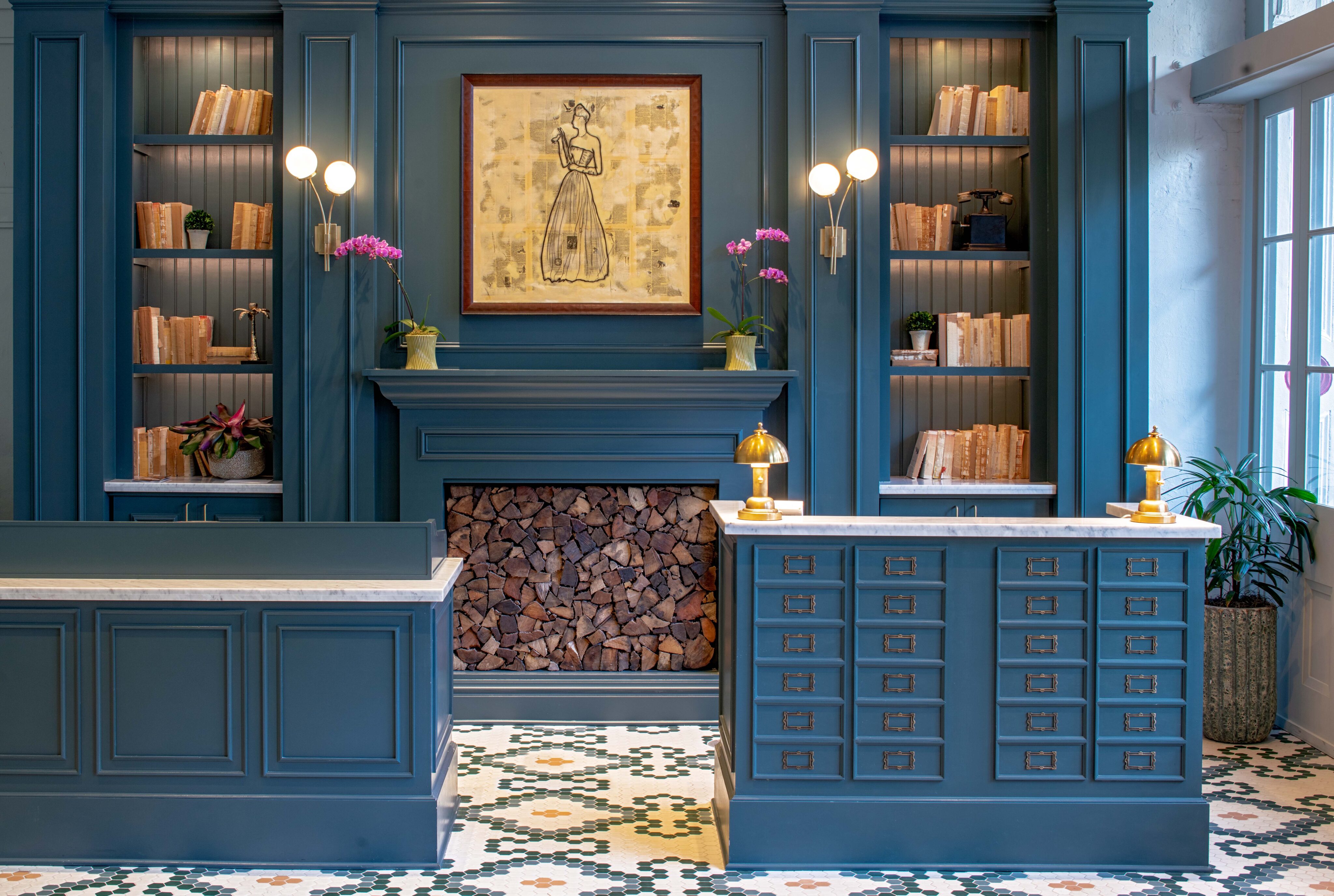 A blue-paneled room with built-in shelves, a fireplace stacked with logs, two desks with brass lamps, and a framed artwork above the mantel.