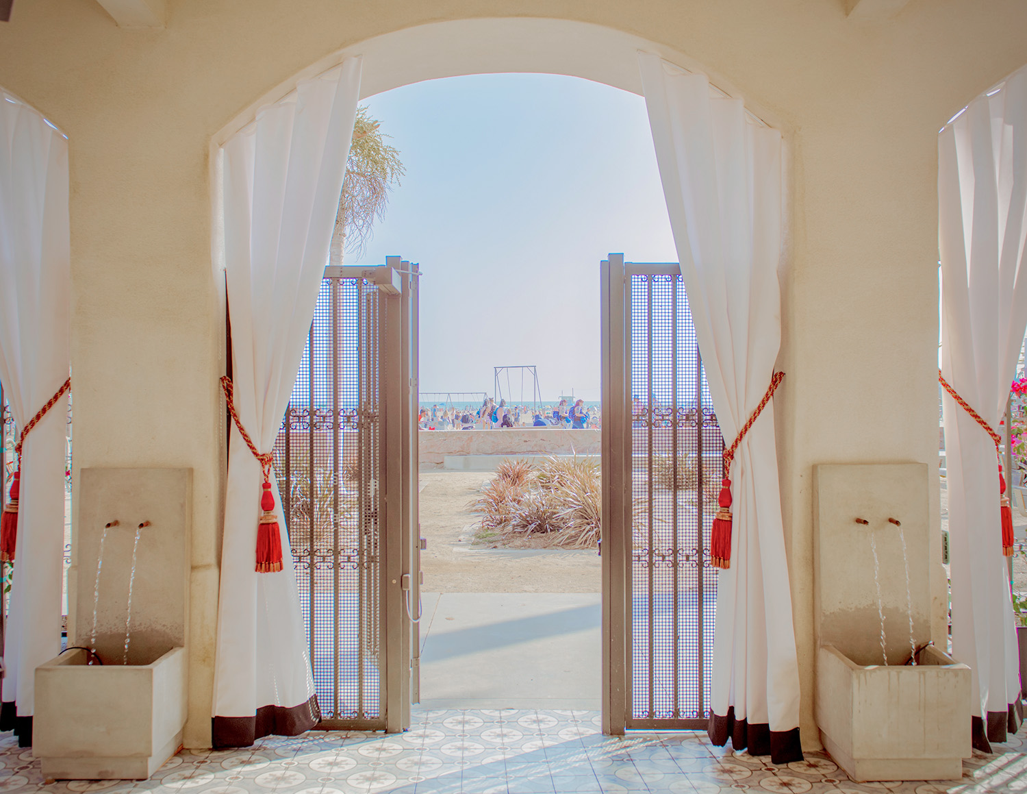 Open double doors with metal gates and white curtains reveal a beach scene with sand, people, and a swing set; two wall fountains are visible in the foreground.