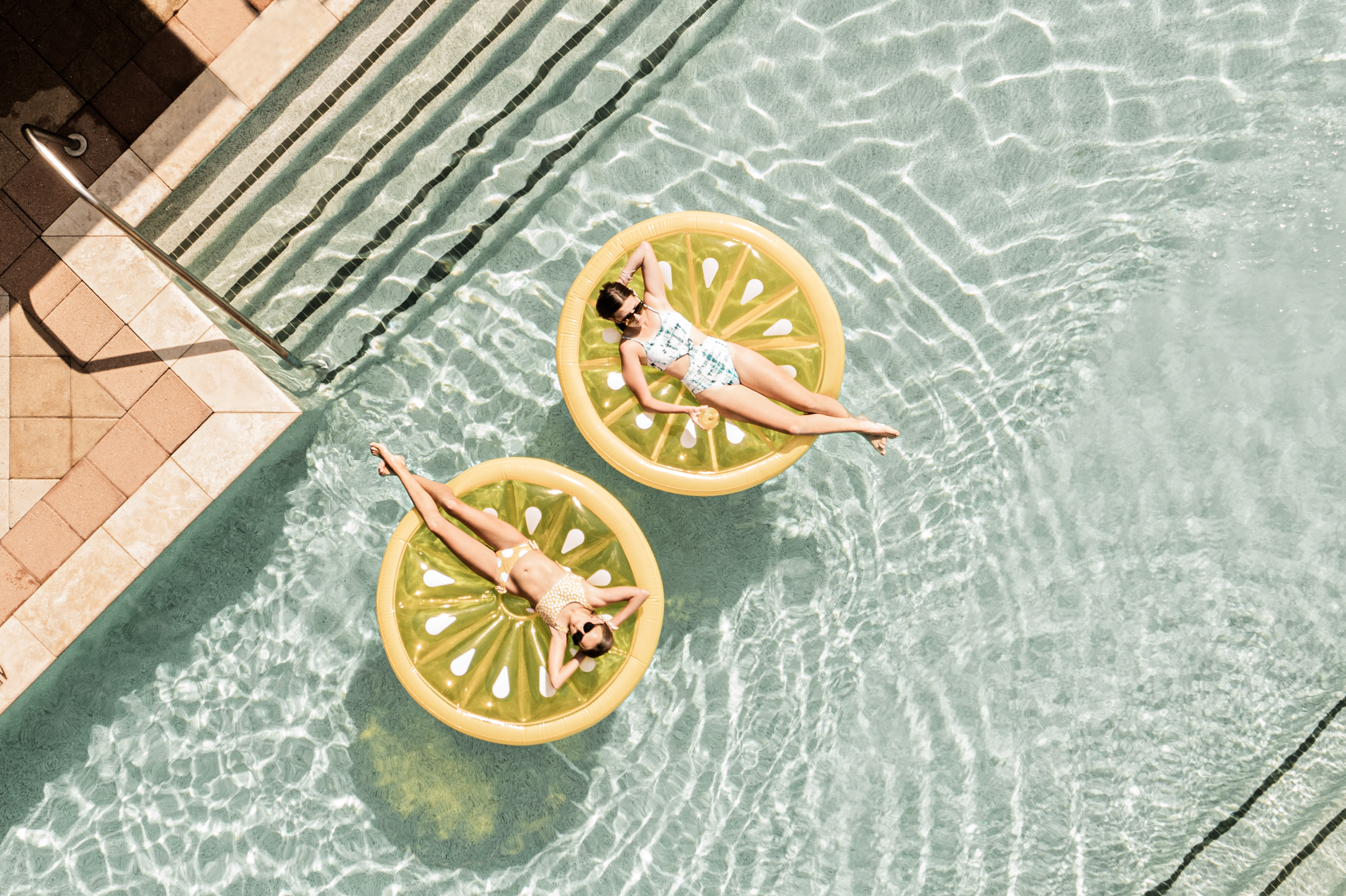 Two people relax on yellow inflatable pool floats shaped like lemon slices in a clear swimming pool near poolside steps.