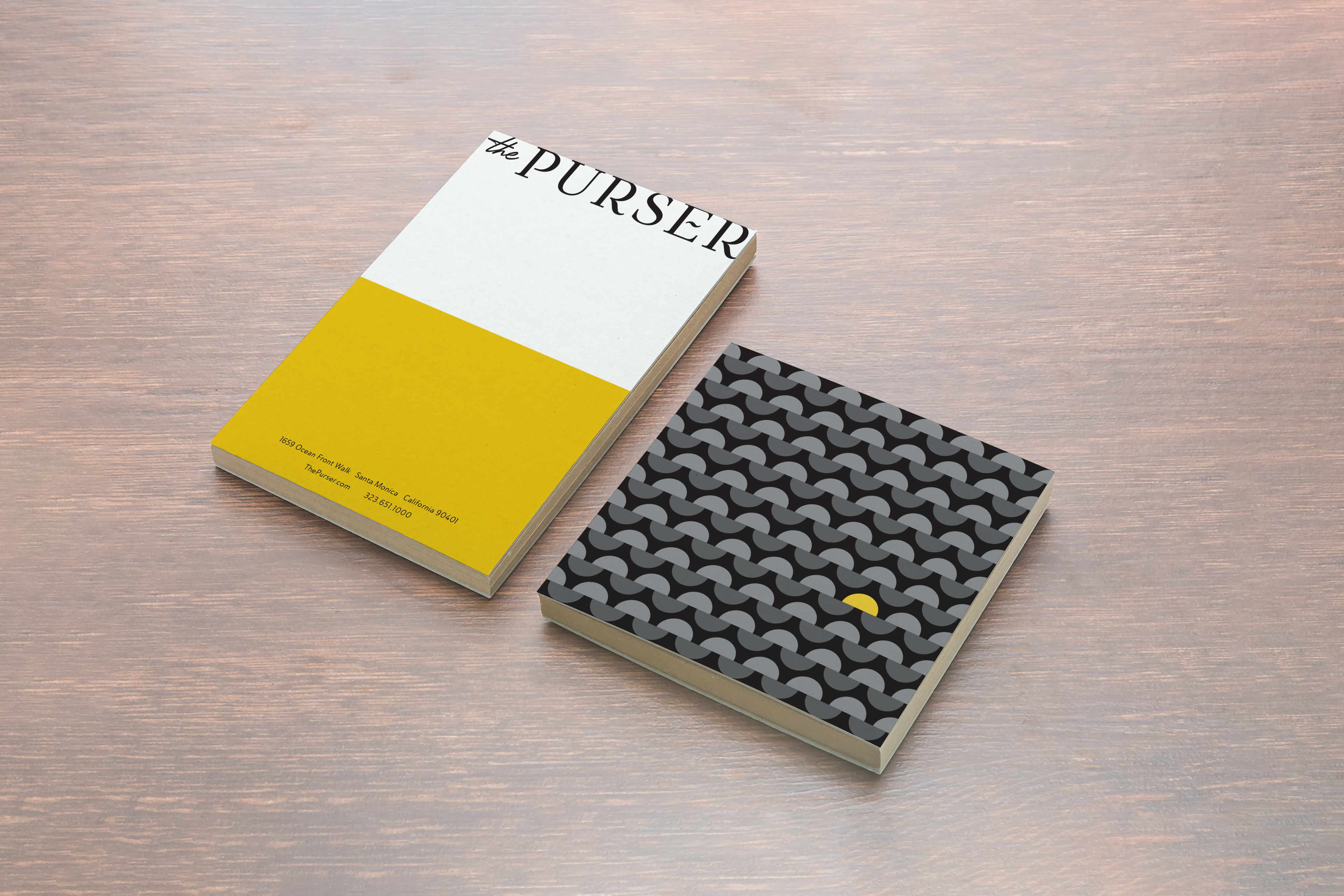 Two closed books are lying on a wooden surface; one has a minimalist yellow and white cover titled "The PURSER," and the other has a black cover with a wavy pattern and a single yellow dot.