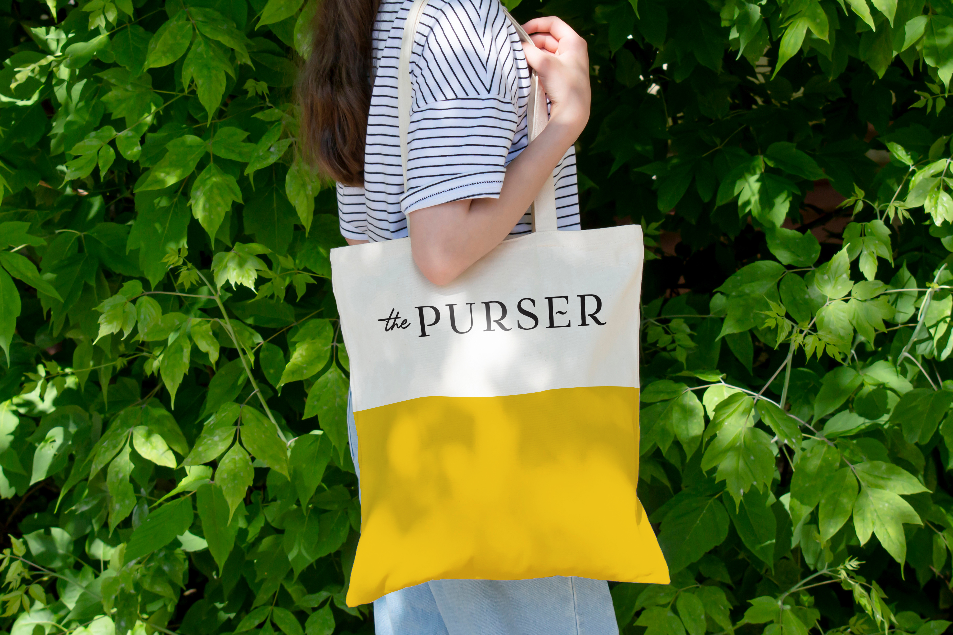 A person wearing a striped shirt holds a yellow and white tote bag labeled "the PURSER" in front of leafy green plants.