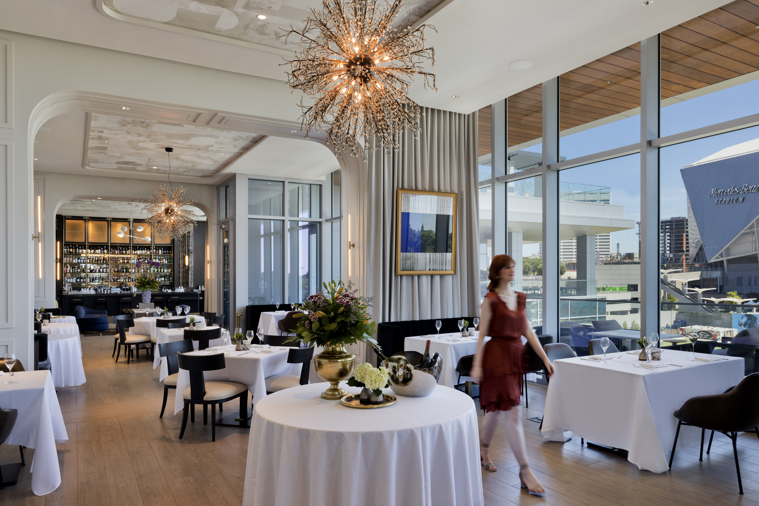 Elegant restaurant interior with white tablecloths, modern chandeliers, large windows, and a woman in a red dress walking past tables.