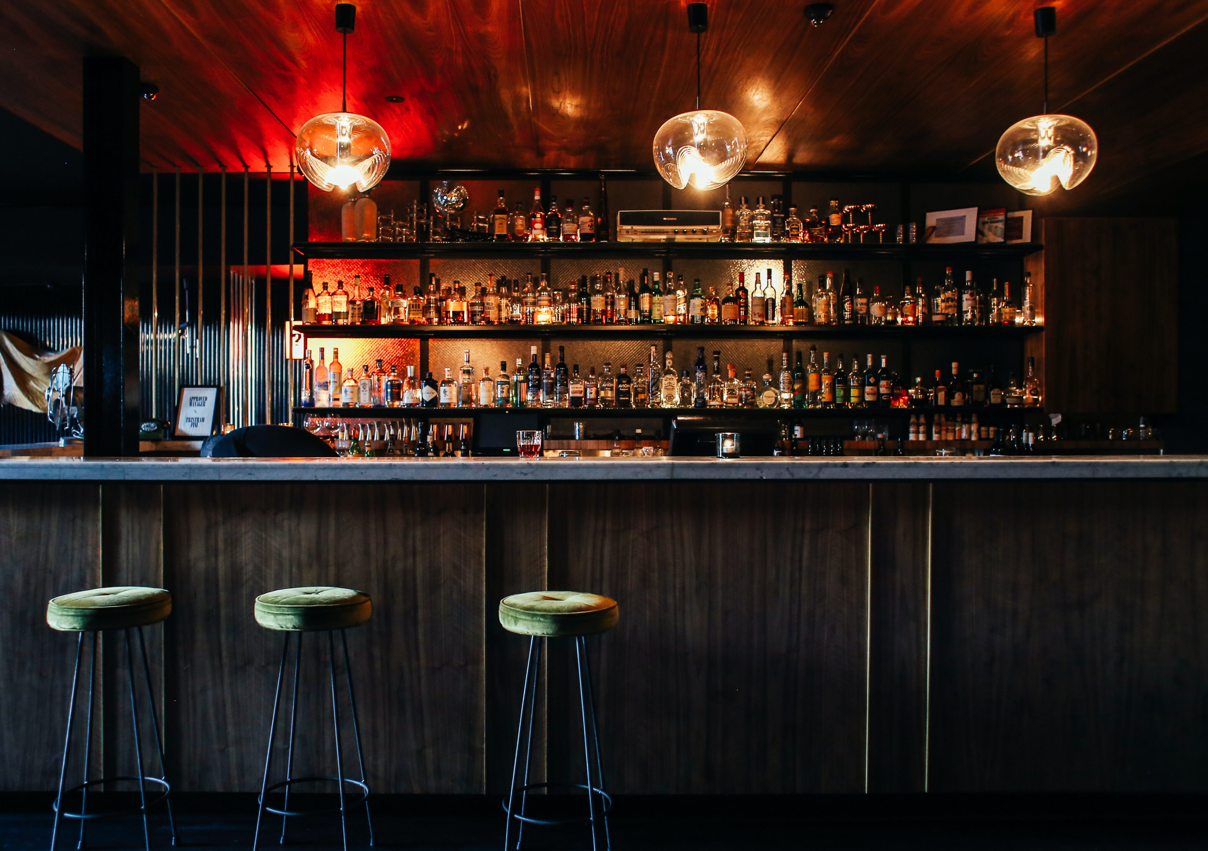 A modern bar interior with three green barstools, a marble countertop, and shelves stocked with various bottles of alcohol, lit by three hanging round lights.