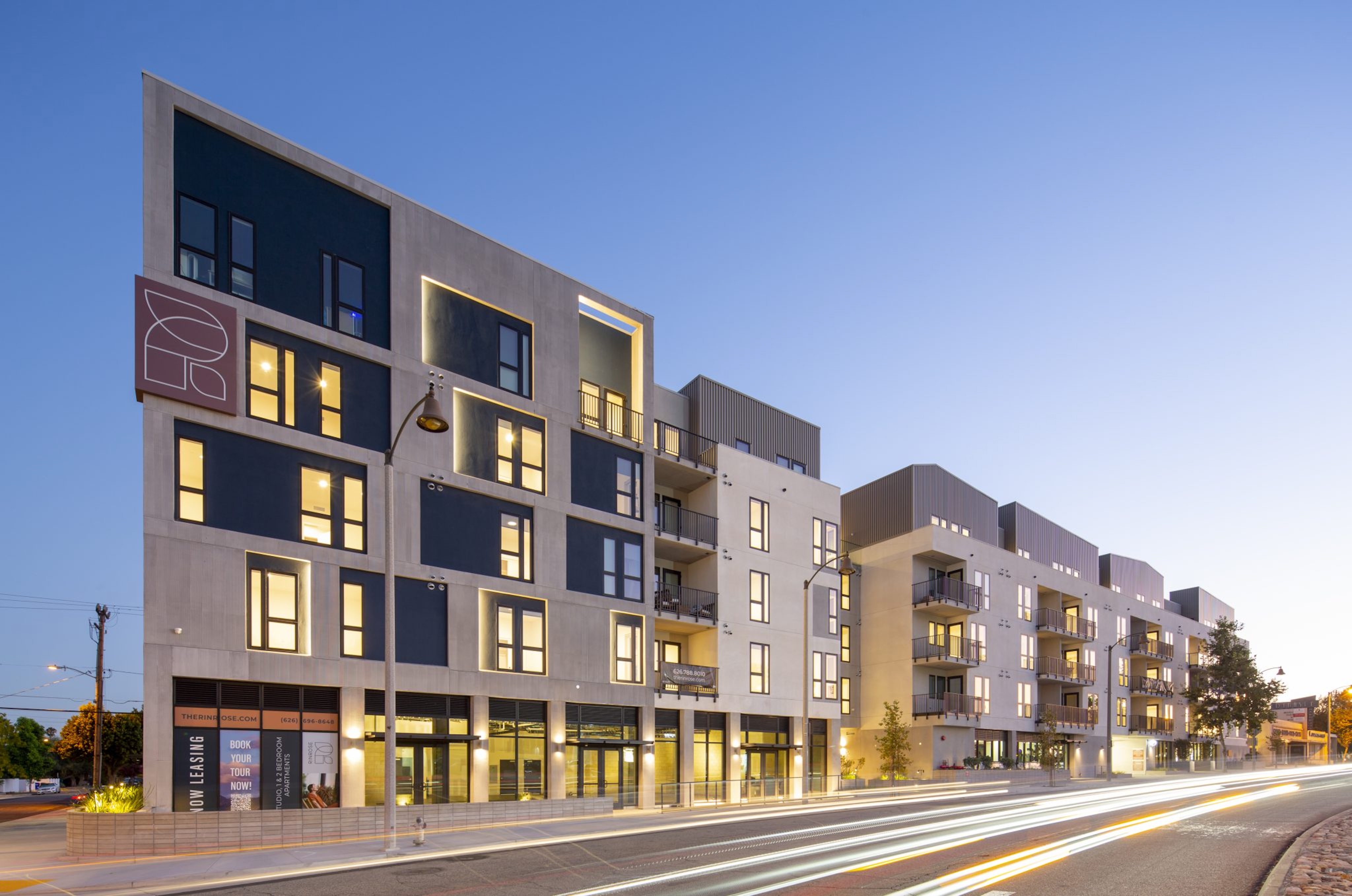 Modern multi-story apartment building with large windows and balconies, illuminated at dusk, with light trails from passing vehicles on the street in front.