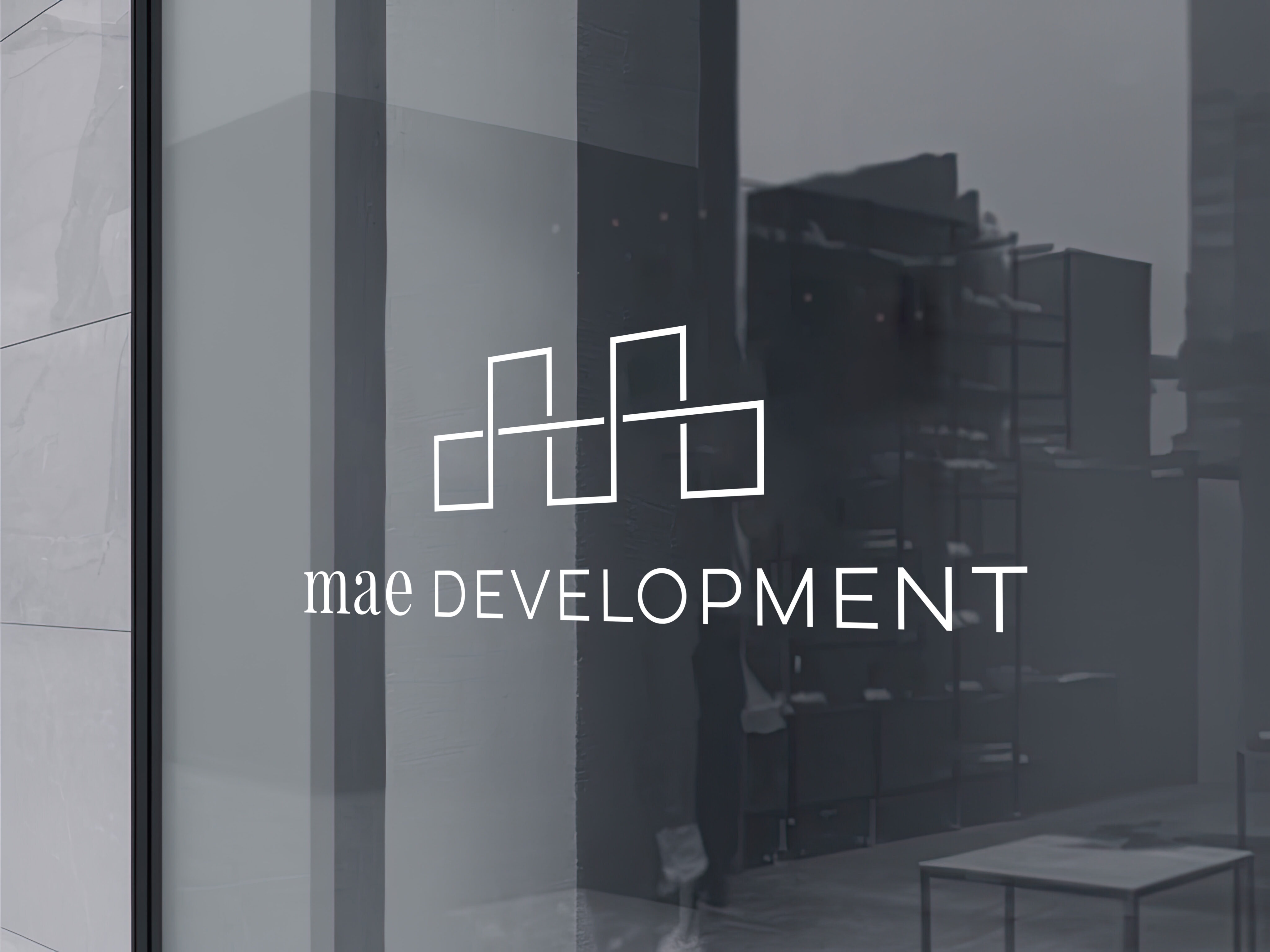 Logo for "mae DEVELOPMENT" displayed on a glass window, featuring a minimalist geometric design above the company name.