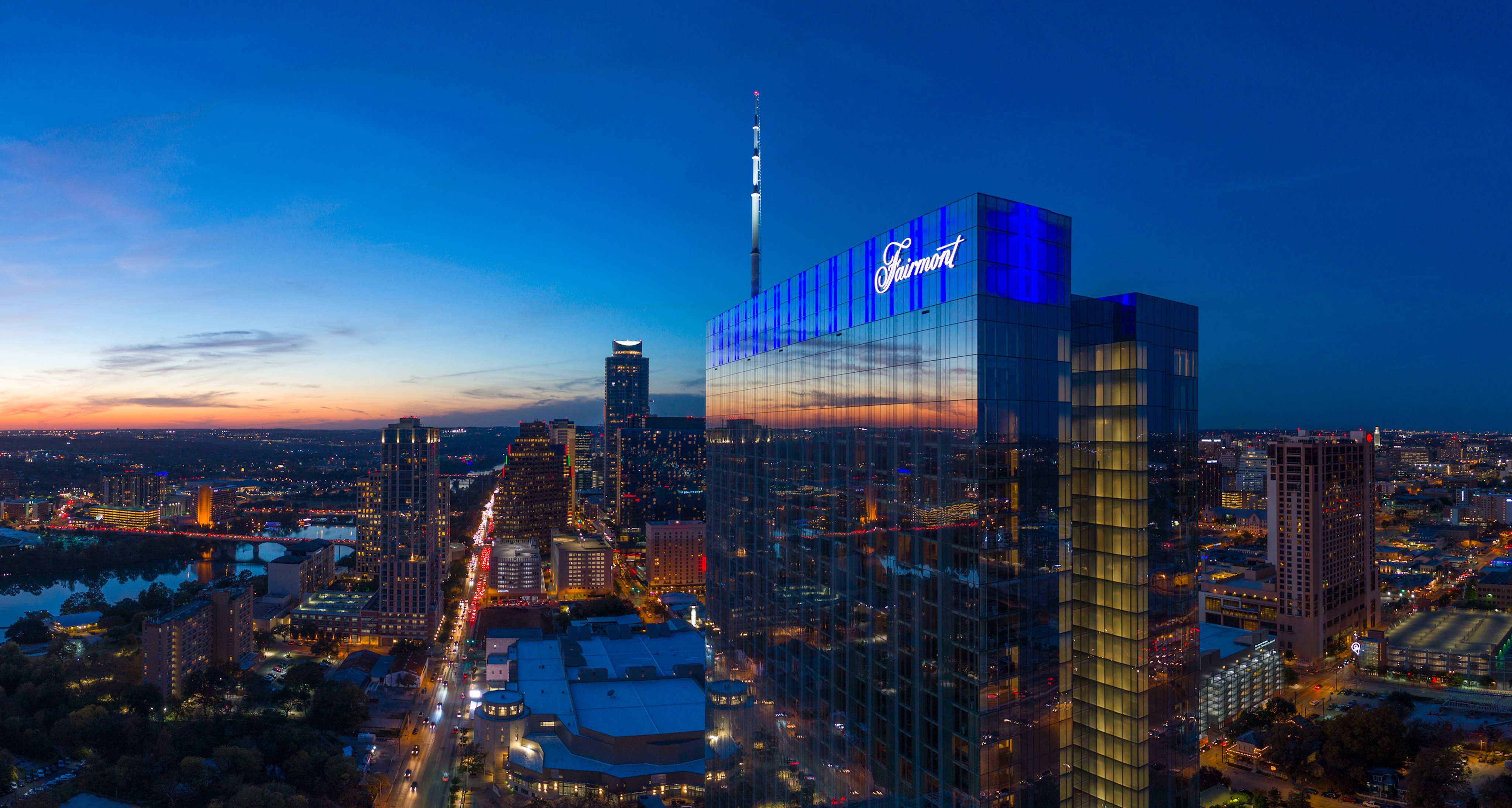 A tall glass building with the Fairmont logo is lit up at dusk, overlooking a cityscape with streets and buildings in downtown Austin, Texas.