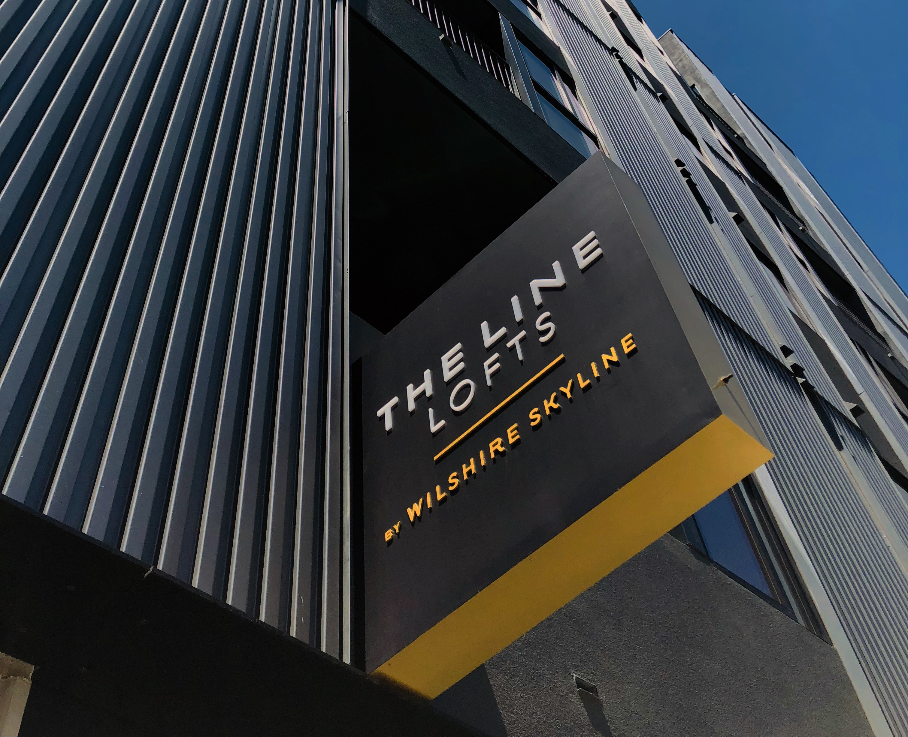 A modern building facade with a black and yellow sign reading “The Line Lofts by Wilshire Skyline” in bold, uppercase letters.
