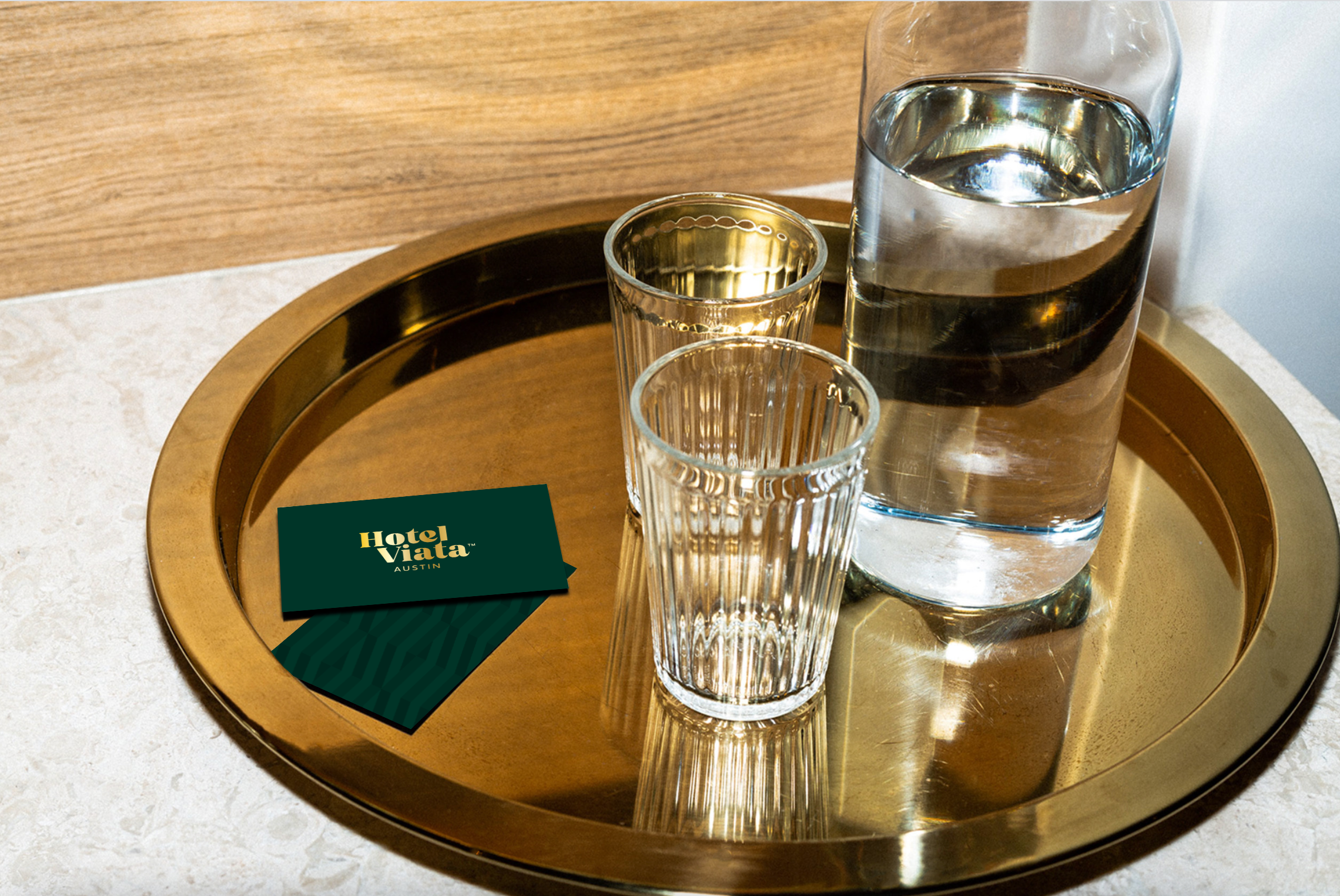 A glass water bottle, two empty ribbed glasses, and two green hotel cards on a round gold tray placed on a beige surface.
