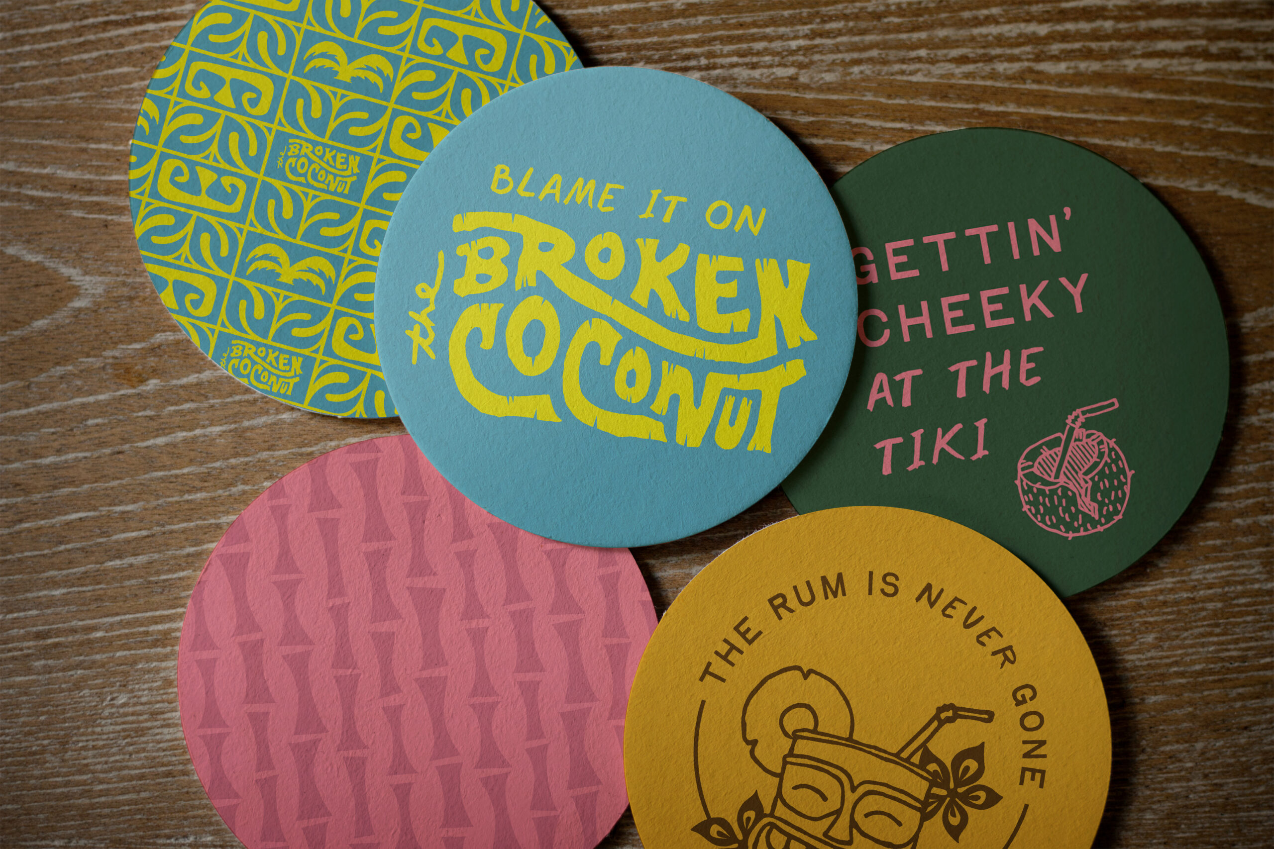 Five colorful drink coasters with playful designs and text, including phrases like "Blame it on the Broken Coconut" and "Gettin' Cheeky at the Tiki," on a wooden surface.