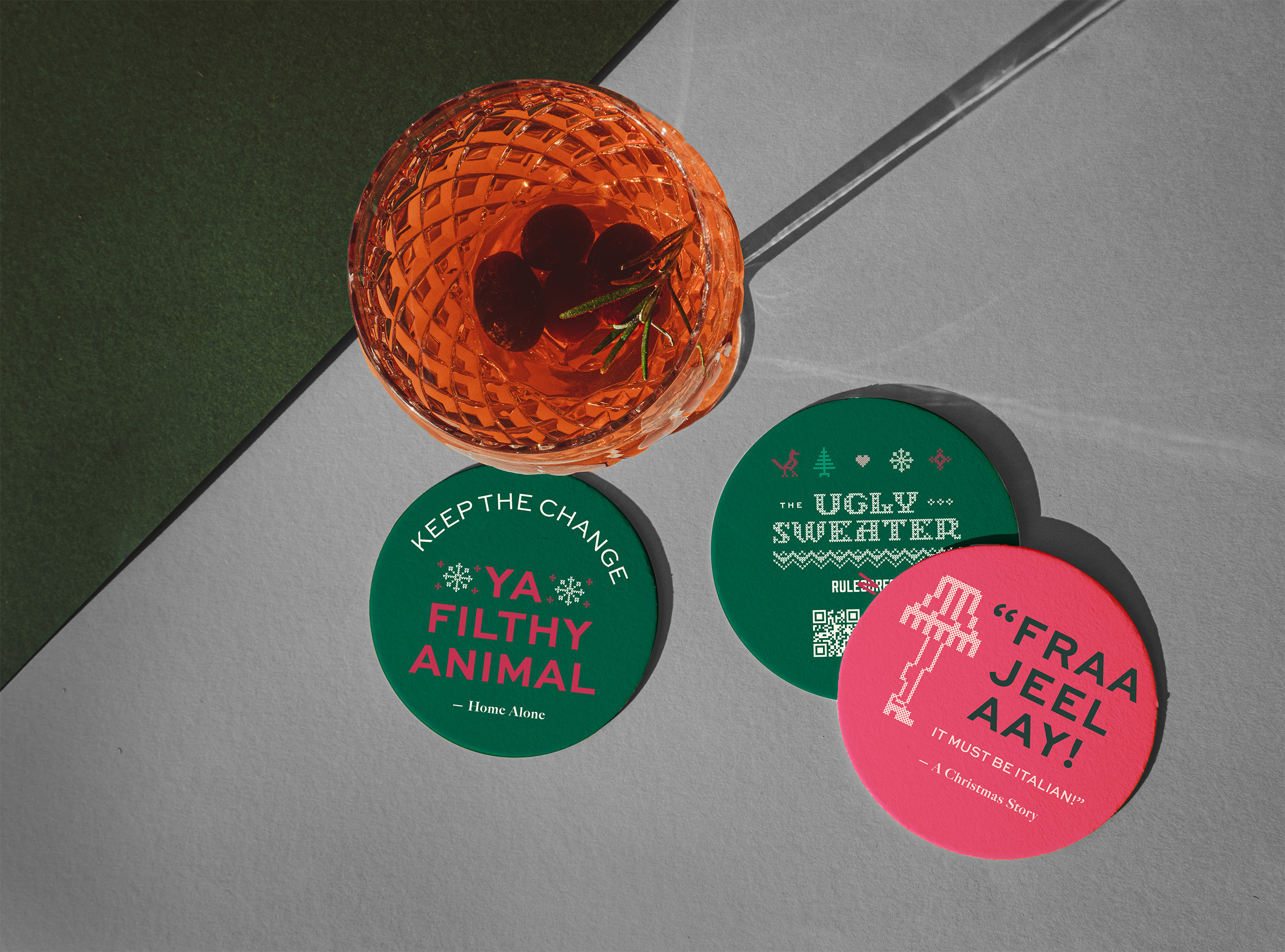 A cocktail with cherries sits beside three colorful coasters featuring festive text and holiday-themed quotes on a geometric surface.