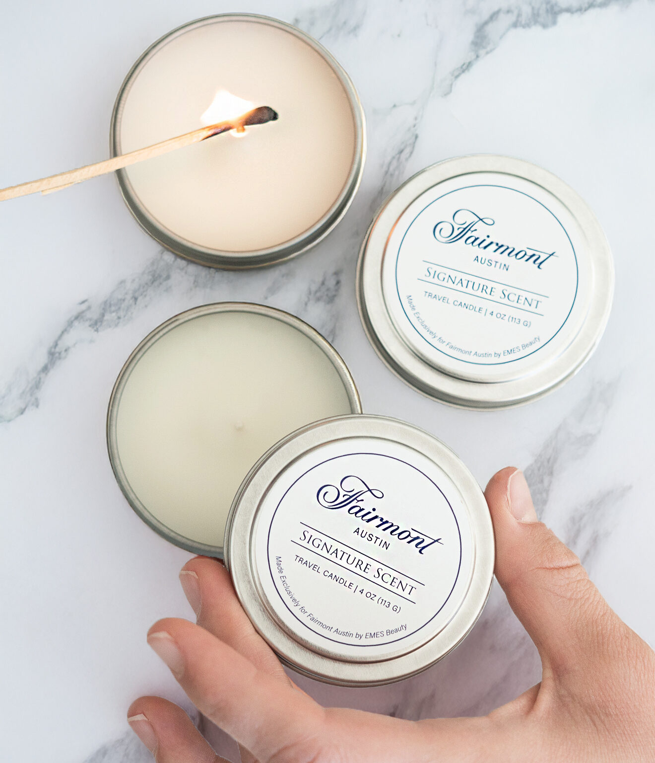 A hand holds a tin labeled "Fairmont Austin Signature Scent" next to two similar candles, one of which is being lit with a wooden stick on a marble surface.