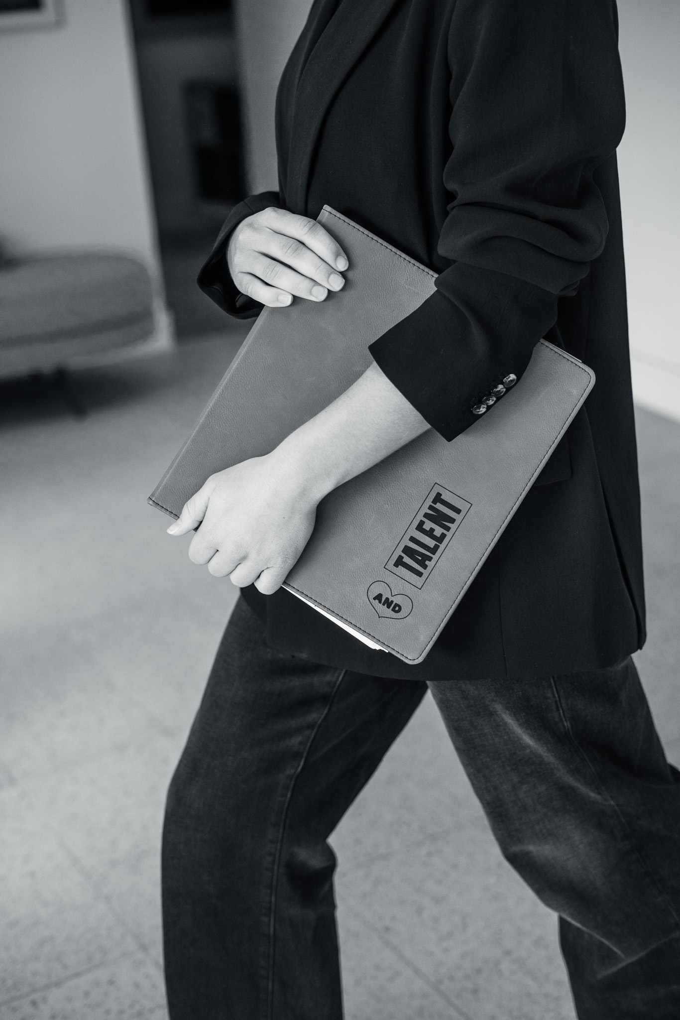 Person in a blazer and jeans holding a large folder labeled "TALENT" while walking indoors.